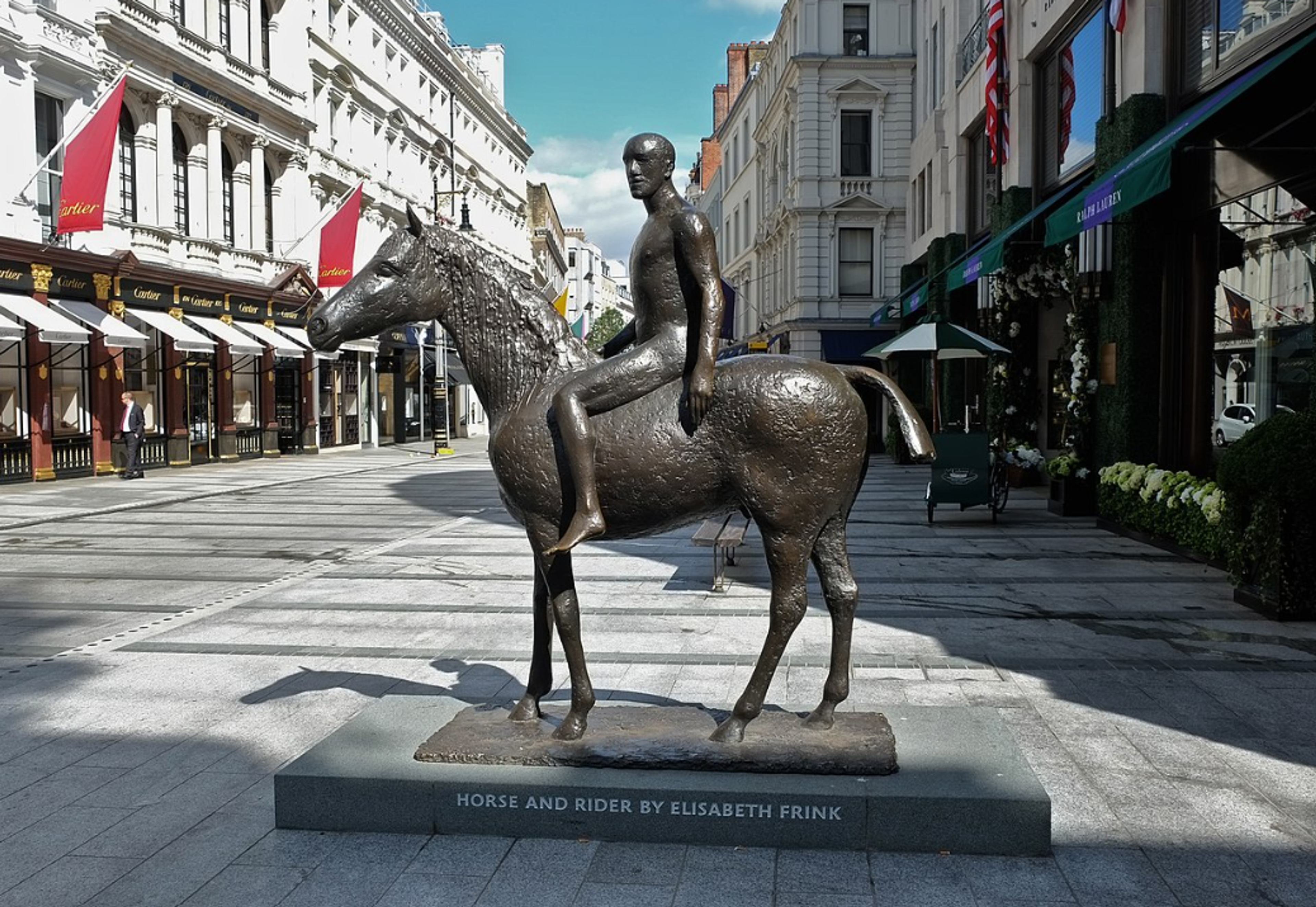 A bronze sculpture of a male figure riding a horse. The horse appears to be stationary, with just his back left leg forward. The rider holds onto the horse’s mane with his right hand, leaning his left back against the horse’s flank. He looks over to the left. The finish of the sculpture is very textured, with small areas of shiny smoothness.