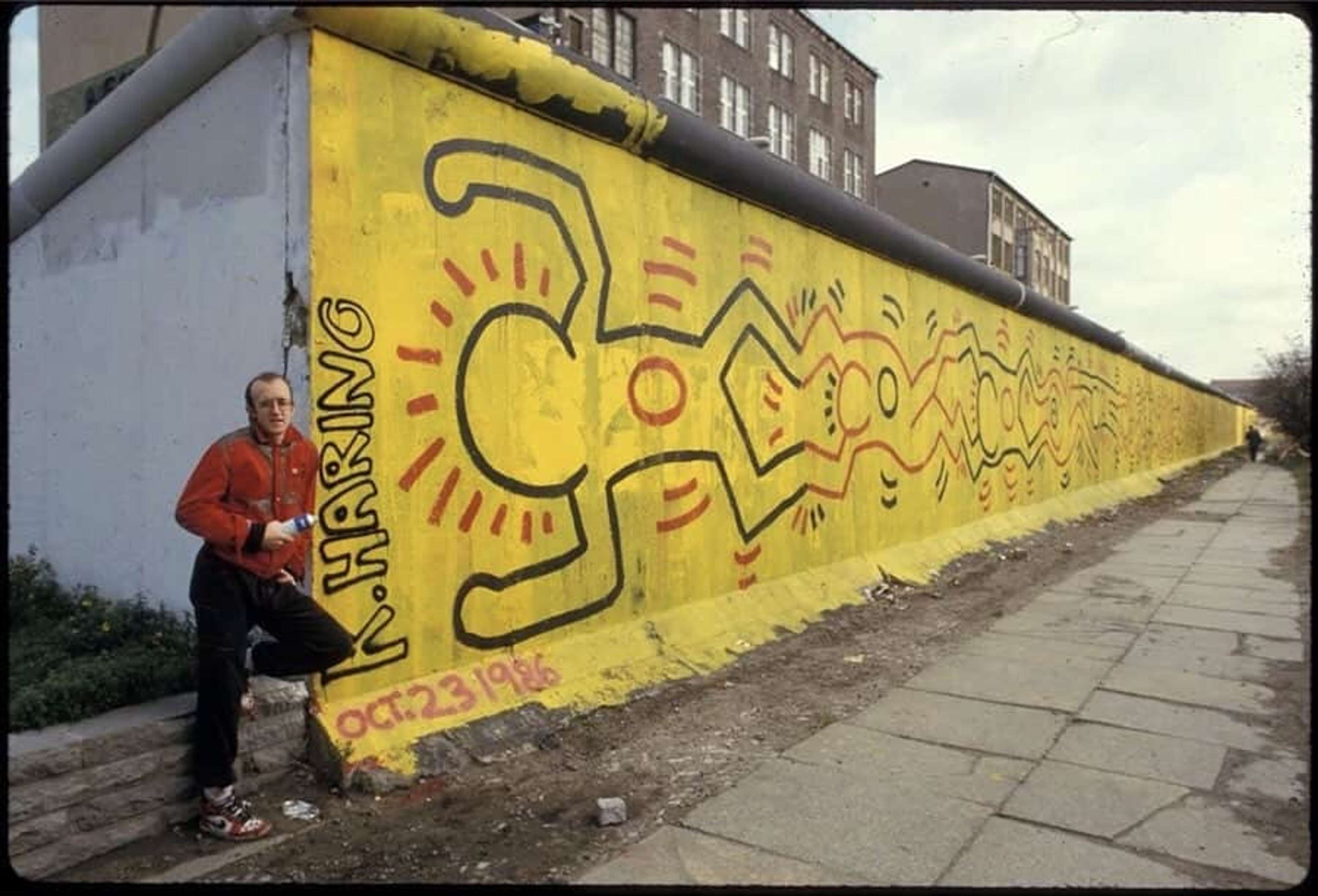 Photographie de l'artiste Keith Haring debout devant sa fresque murale sur le mur de Berlin. La fresque représente plusieurs figures reliées entre elles en rouge et noir, sur un fond jaune.