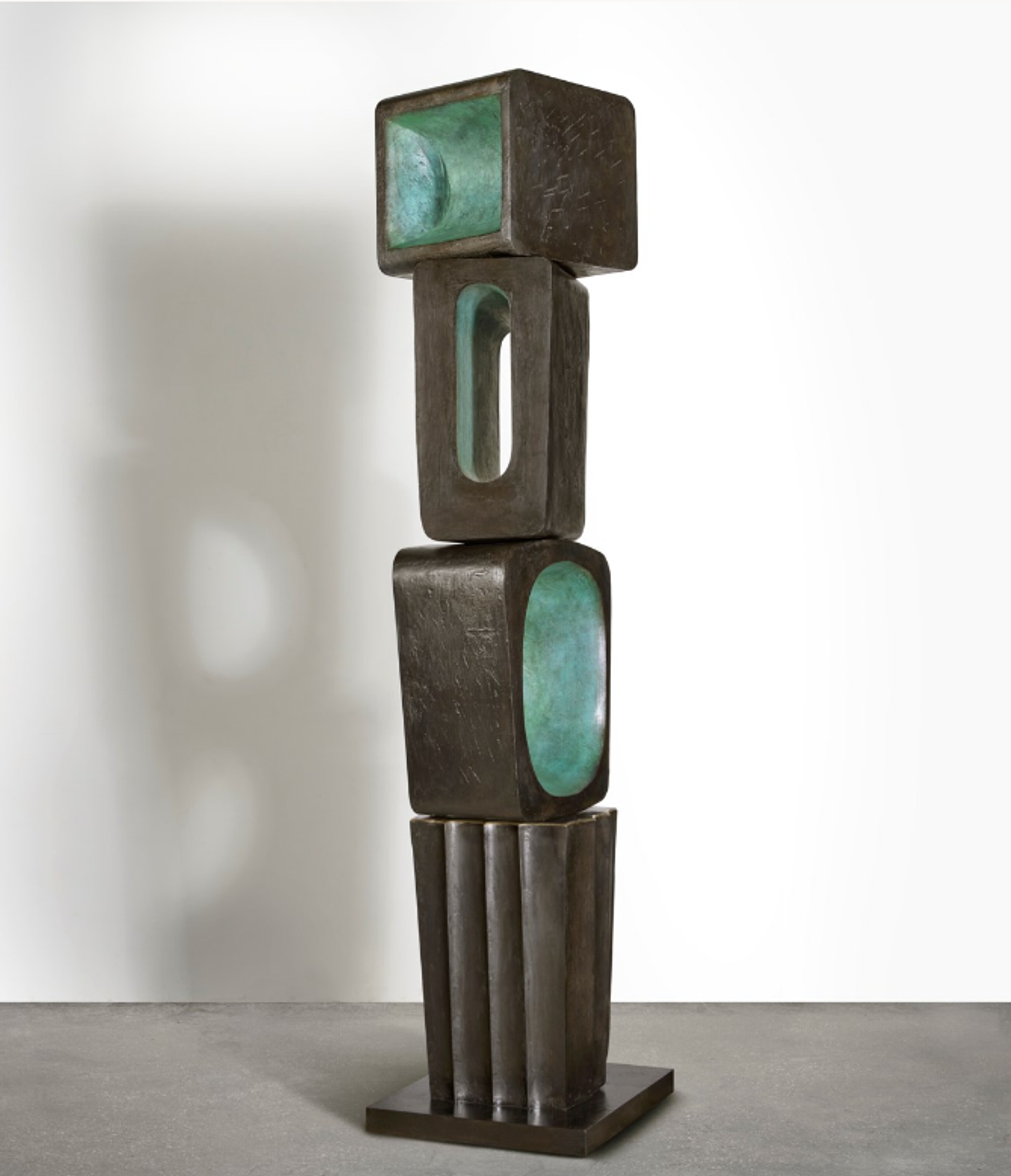 A vertical bronze sculpture of stacked geometric elements resembling a totem pole. The piece features rectangular forms with hollowed sections and interior spaces that reveal turquoise-green colouration. The exterior surface has a dark patina while the recessed areas provide contrasting colours, creating a pattern of solid forms and voids.