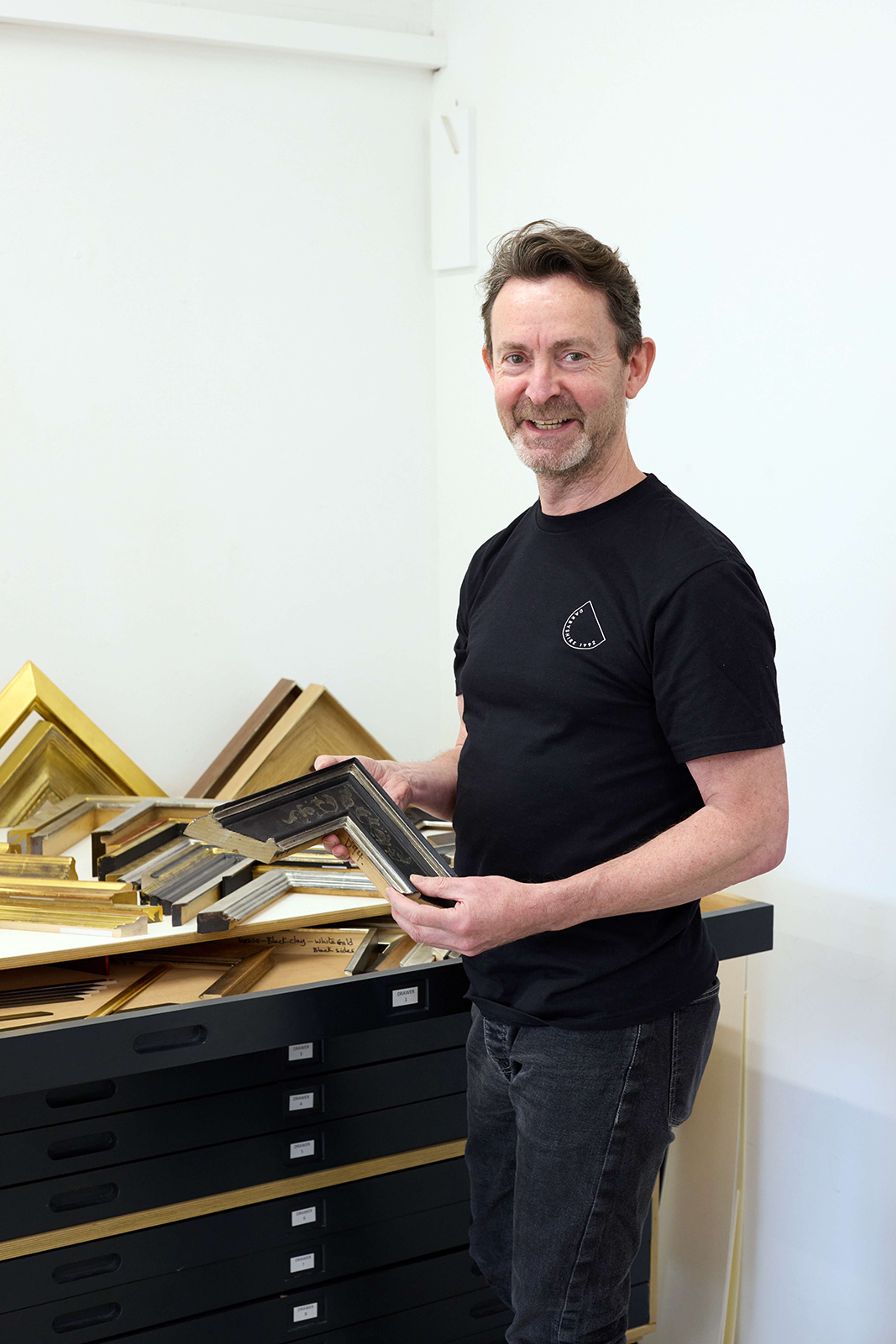 Richard Law, consultant en encadrement chez Darbyshire, se tient à côté d'une table d'échantillons de cadres, tenant un coin de cadre dans un atelier lumineux.
