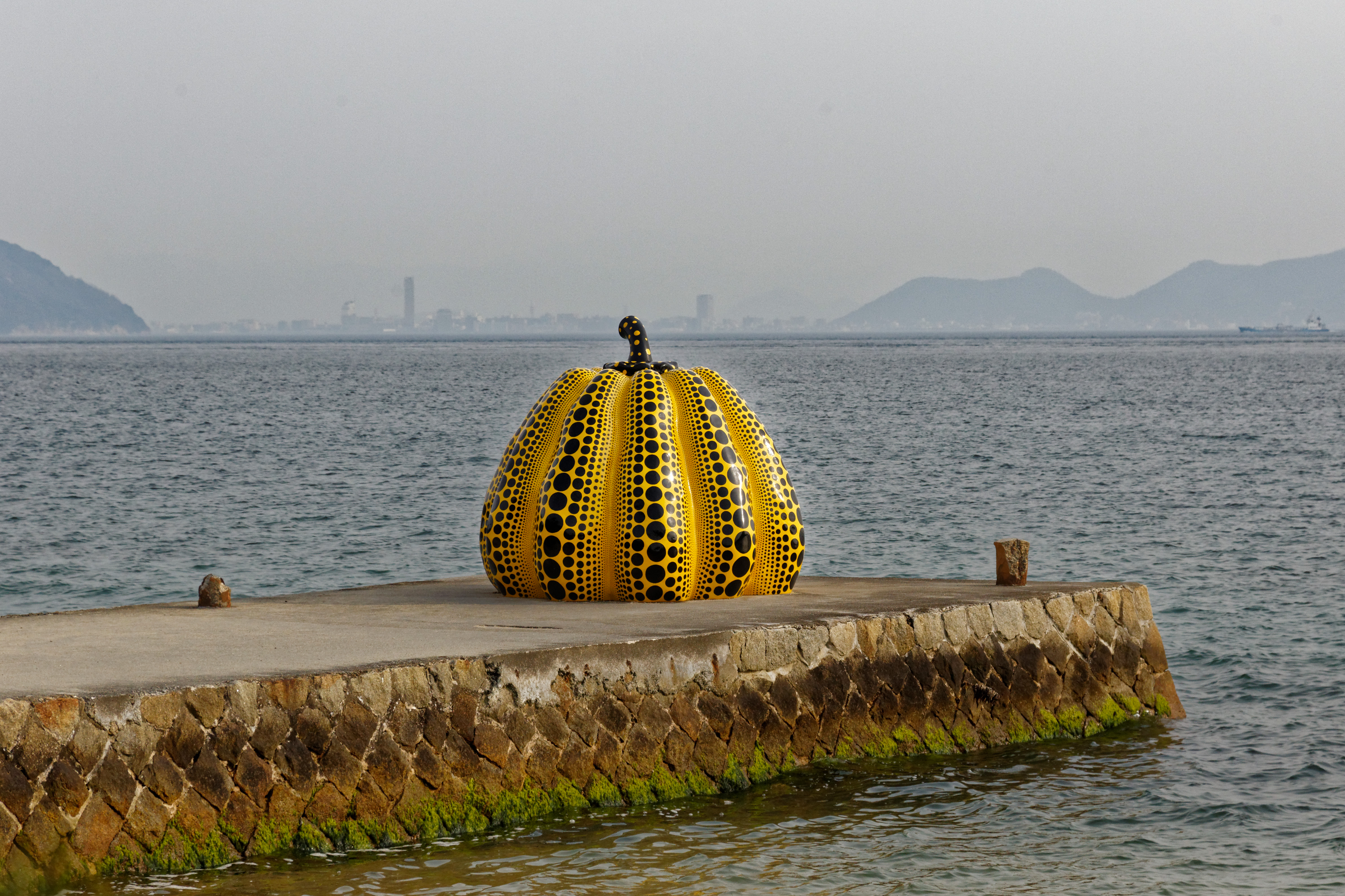 On voit une des sculptures de citrouille géante de Yayoi Kusama sur fond de scène de port intitulée « Harbour Scene ».