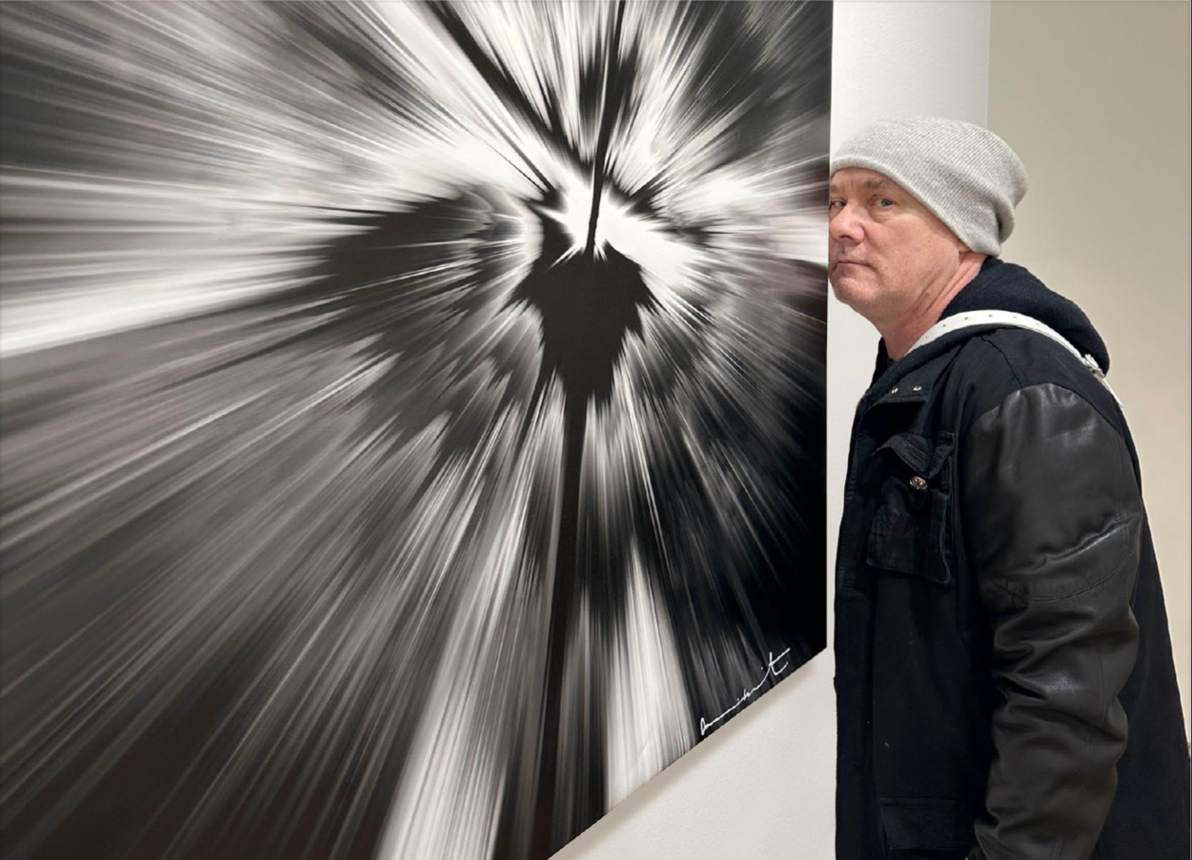L'artiste Damien Hirst, vêtu d'une veste en cuir noir, se tient à côté d'une de ses Spin Paintings. L'œuvre imprimée est au format carré et est composée de noir et blanc, avec la signature de l'artiste dans le coin inférieur droit.