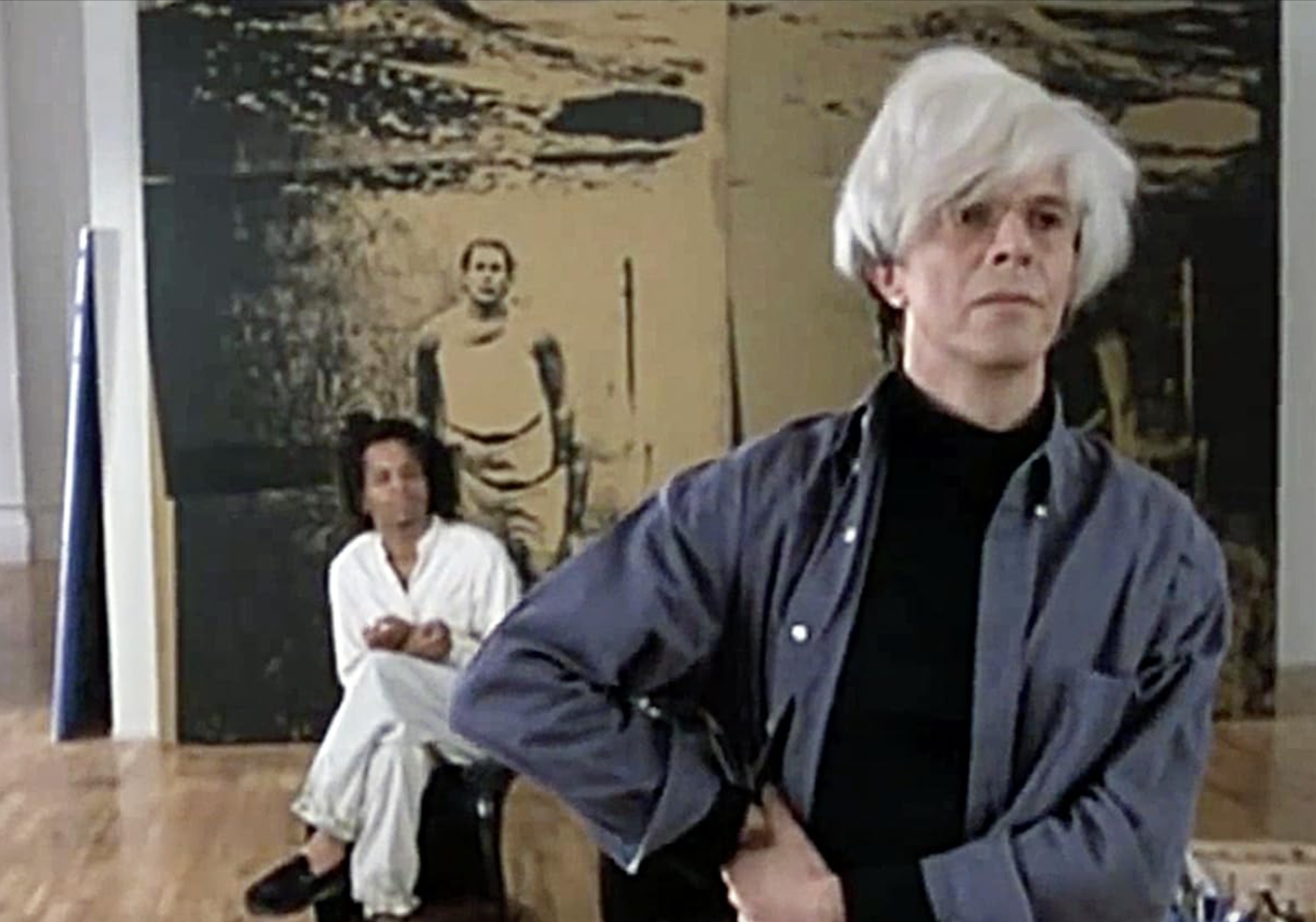 Une capture d'écran du film Basquiat de Julian Schnabel. Elle montre Andy Warhol, interprété par David Bowie, avec Jeffrey Wright dans le rôle de Basquiat à l'arrière-plan.
