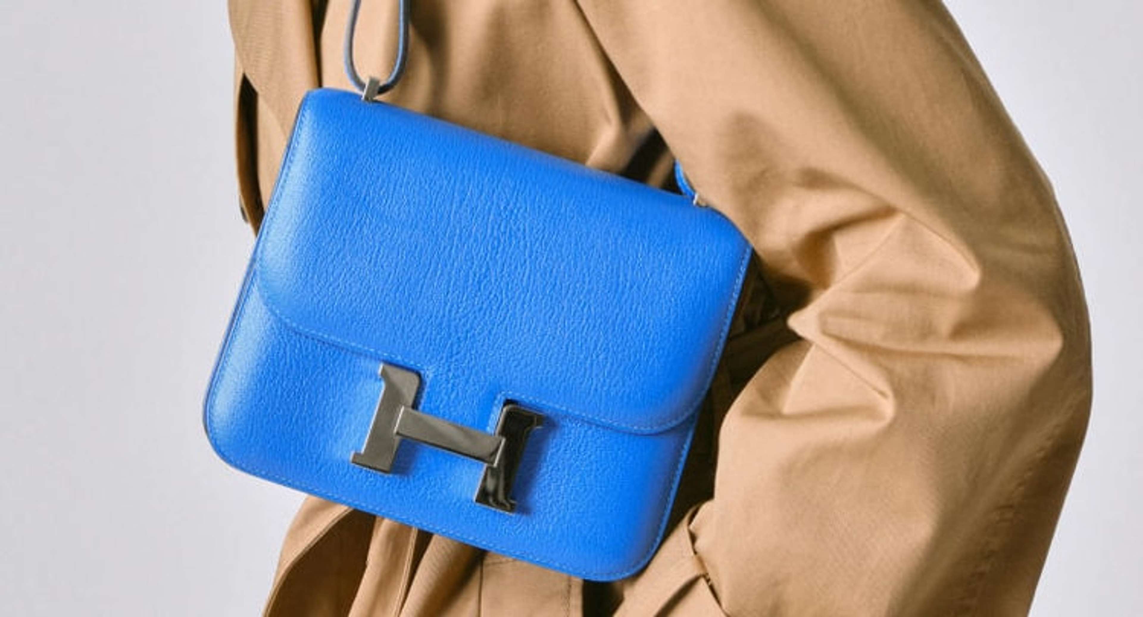 Une image d'une personne vêtue d'un trench-coat, tenant un sac Hermès bleu roi.