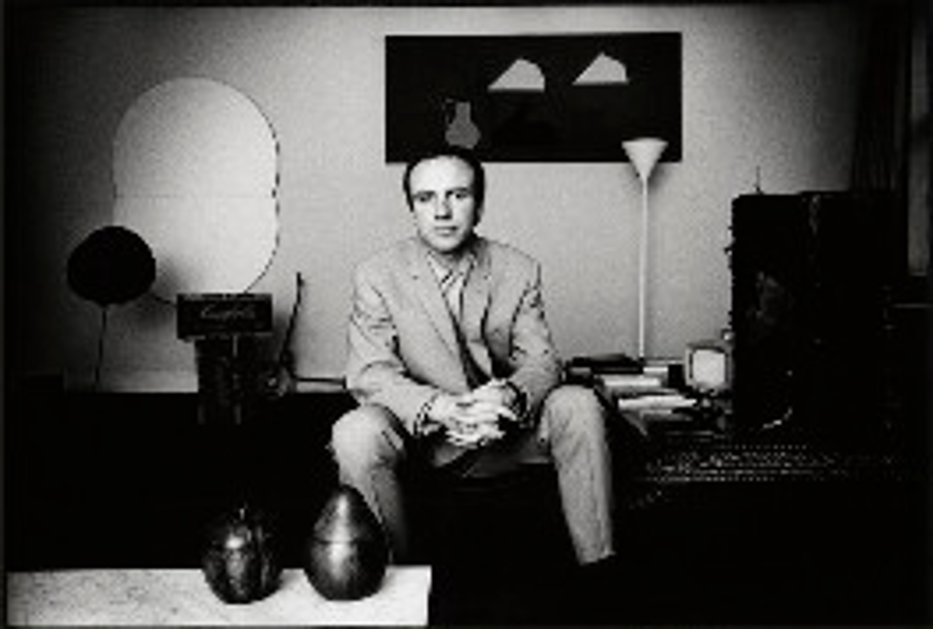Photographie monochrome représentant l'artiste Patrick Caulfield assis sur un banc dans un espace intérieur bien éclairé, regardant directement l'objectif de l'appareil photo.