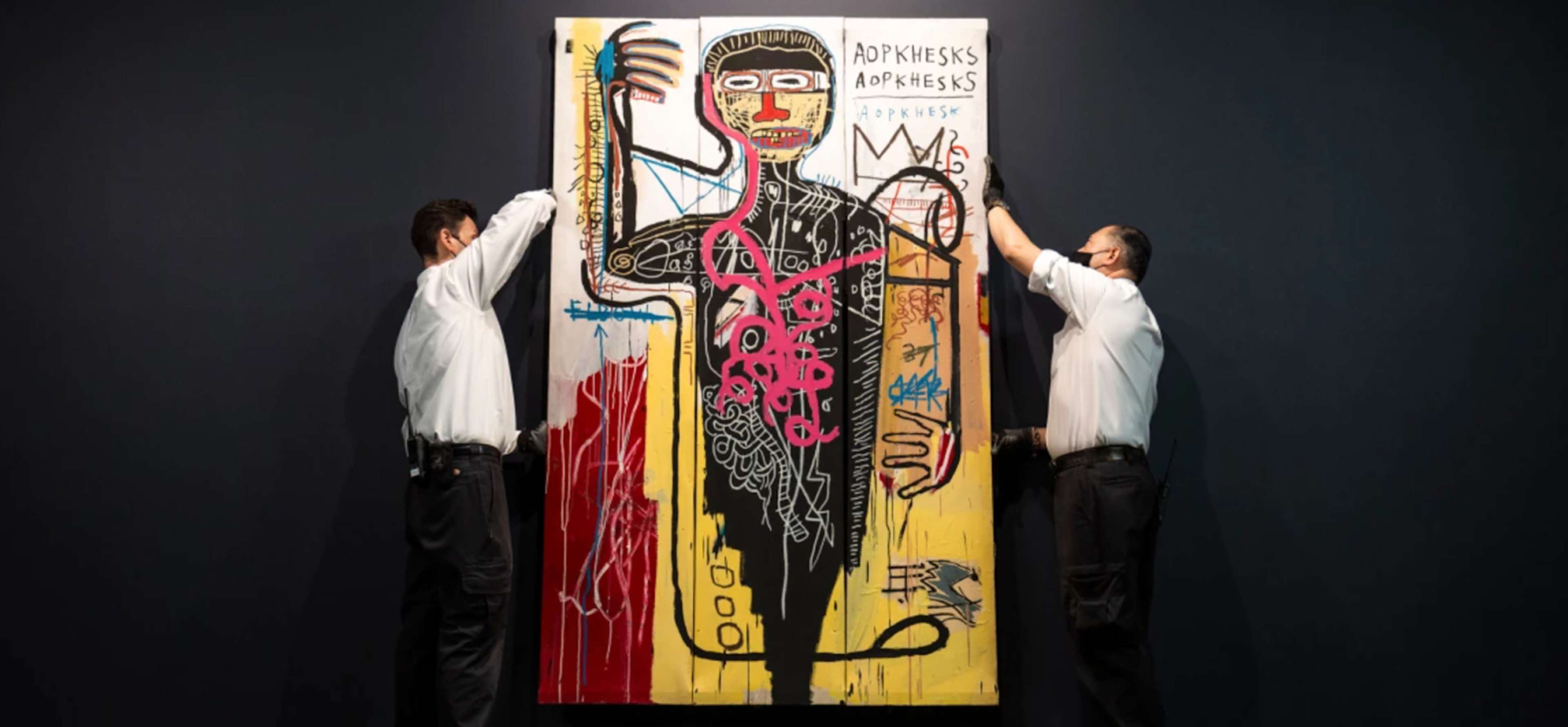 Une image de l'œuvre Versus Medici transportée par deux manutentionnaires d'art chez Sotheby's. Elle montre une figure en noir, entourée des motifs de griffonnages et de couronnes caractéristiques de Basquiat.