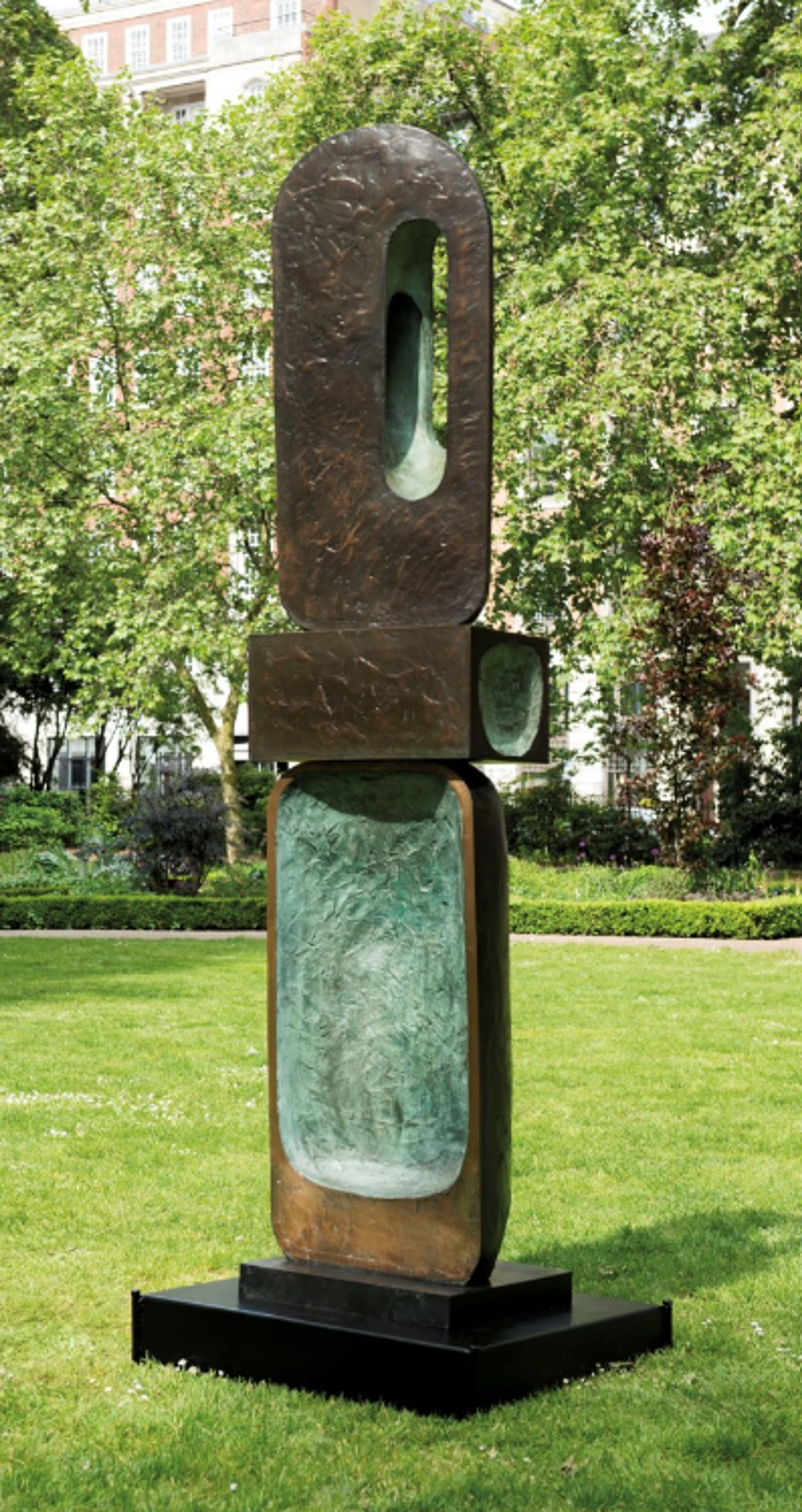 This tall sculpture consists of three stacked geometric elements. The lower piece is wide and thin, with a concave oblong of textured green; the middle piece is a short geometric cuboid; the top piece is a rounded oblong with a smaller version cut out of the right side. The bronze has a rich patina with green and brown tones. 