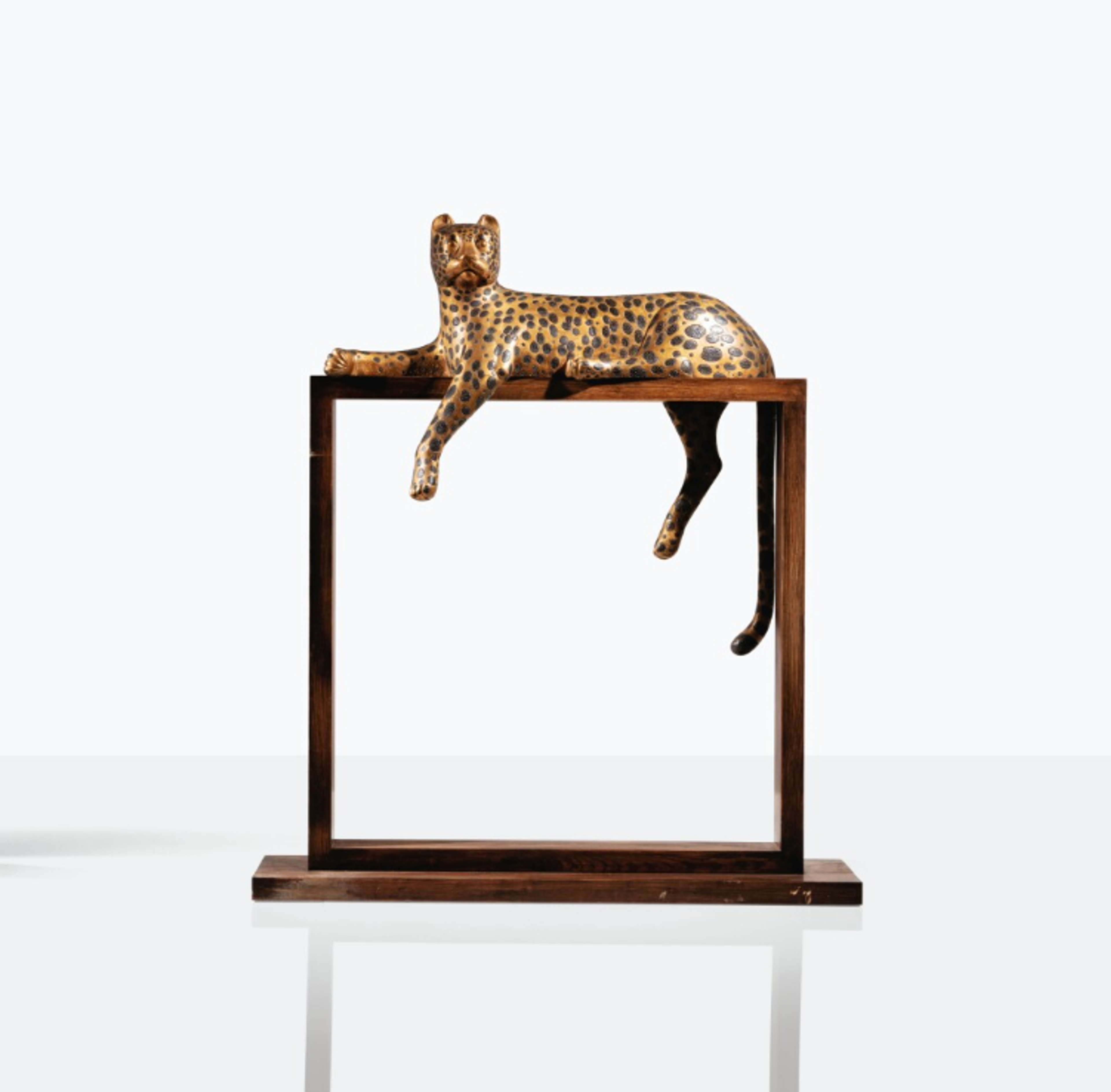 A bronze sculpture of a leopard reclining on a rectangular wooden frame. The sleek feline form appears to be in a relaxed pose, with two legs tucked beside its body and its other two legs and tail hanging down into the frame. The leopard looks towards the viewer.
