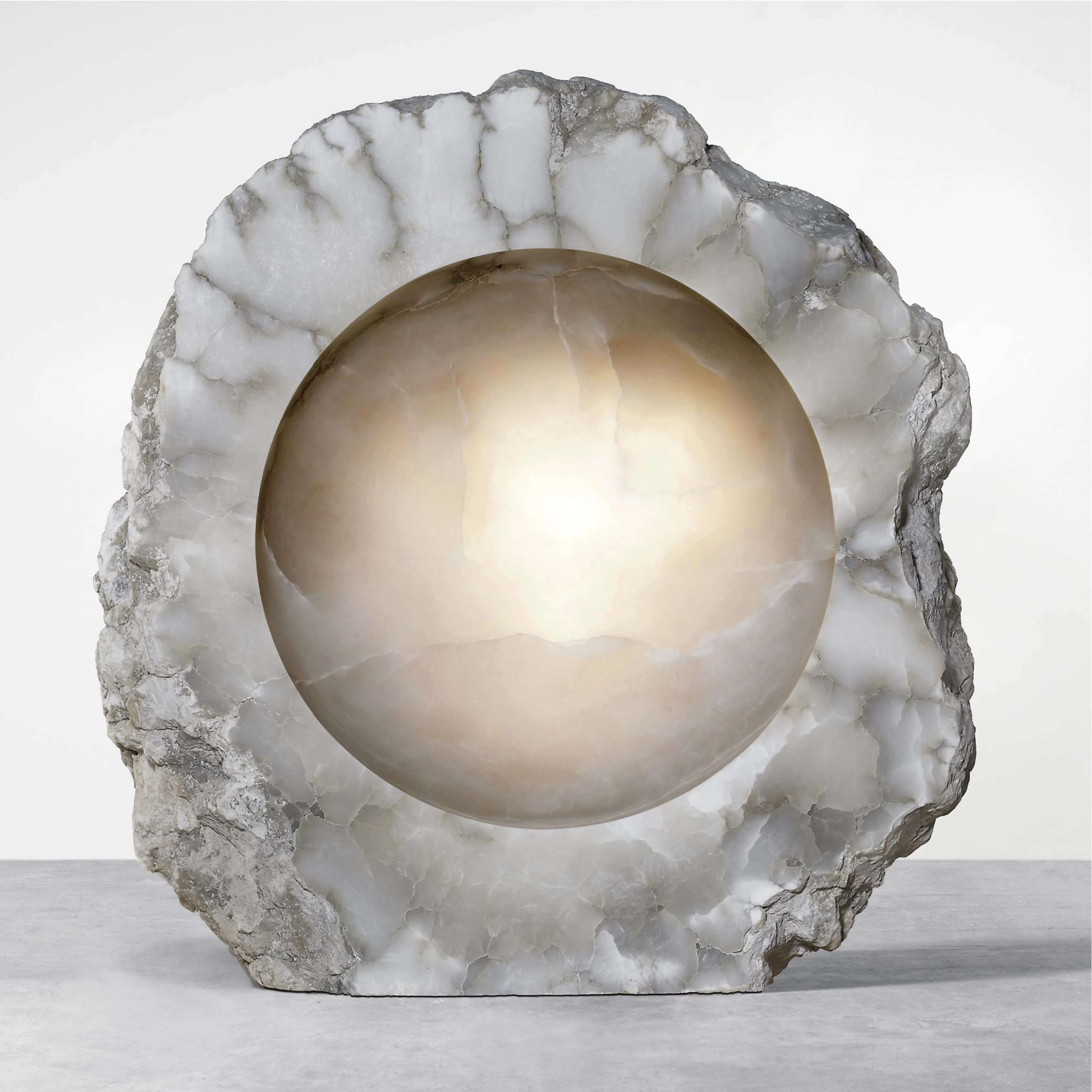 A roughly hewn semi-translucent alabaster rock with dark grey veins. The edges are left jagged, but the vertical face has been polished perfectly smooth. Out of this face, a concave half-sphere has been carved, again with a perfectly smooth surface. The colour of the half-sphere has a warmer tinge.