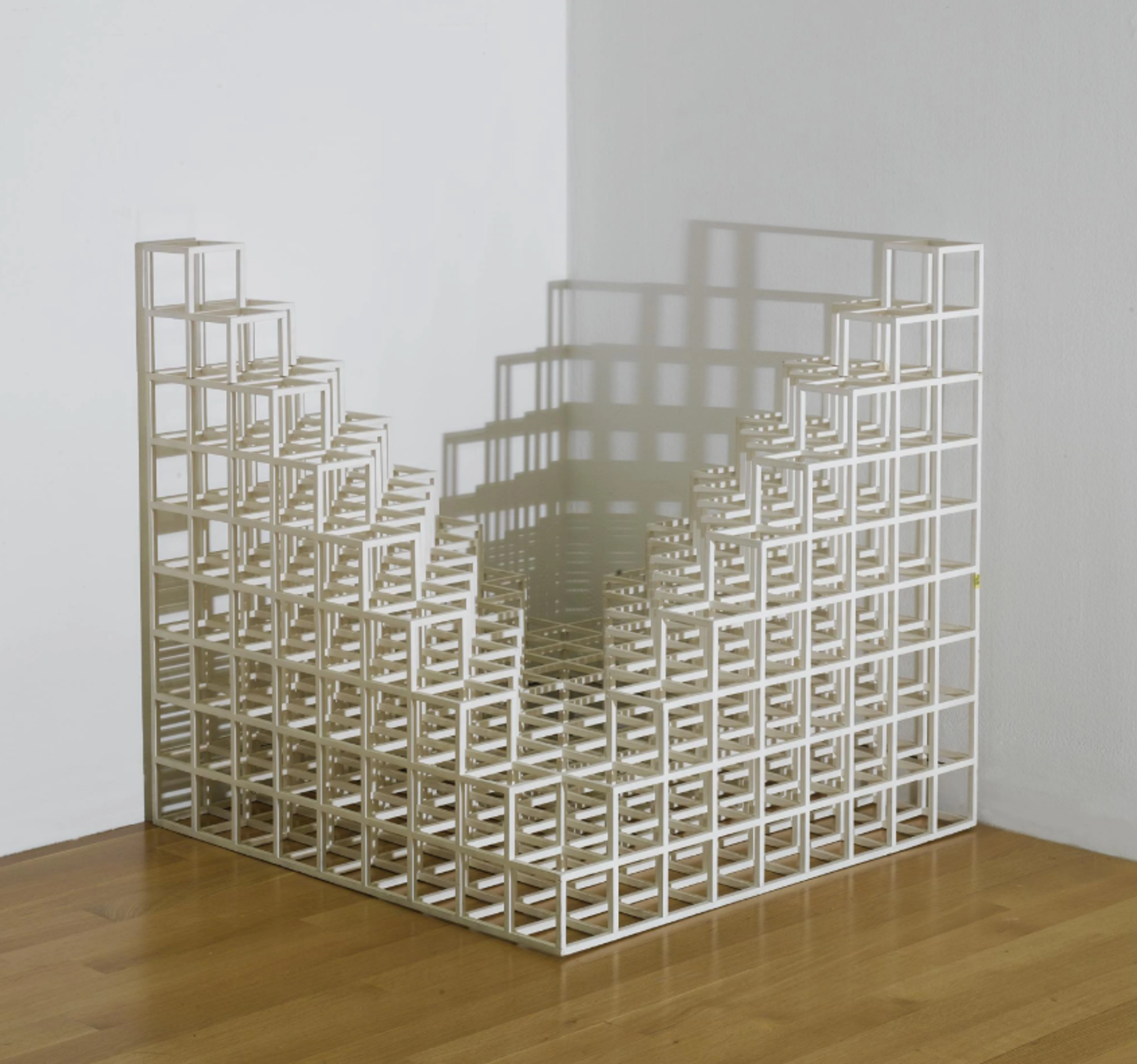 A geometric arrangement of open white cubes set into the corner of a room. The cubes form the two opposite corners of a larger open cube, with two taller graduating stacks sitting opposite each other against the wall. The cubes are only one cube deep on the base and sides.