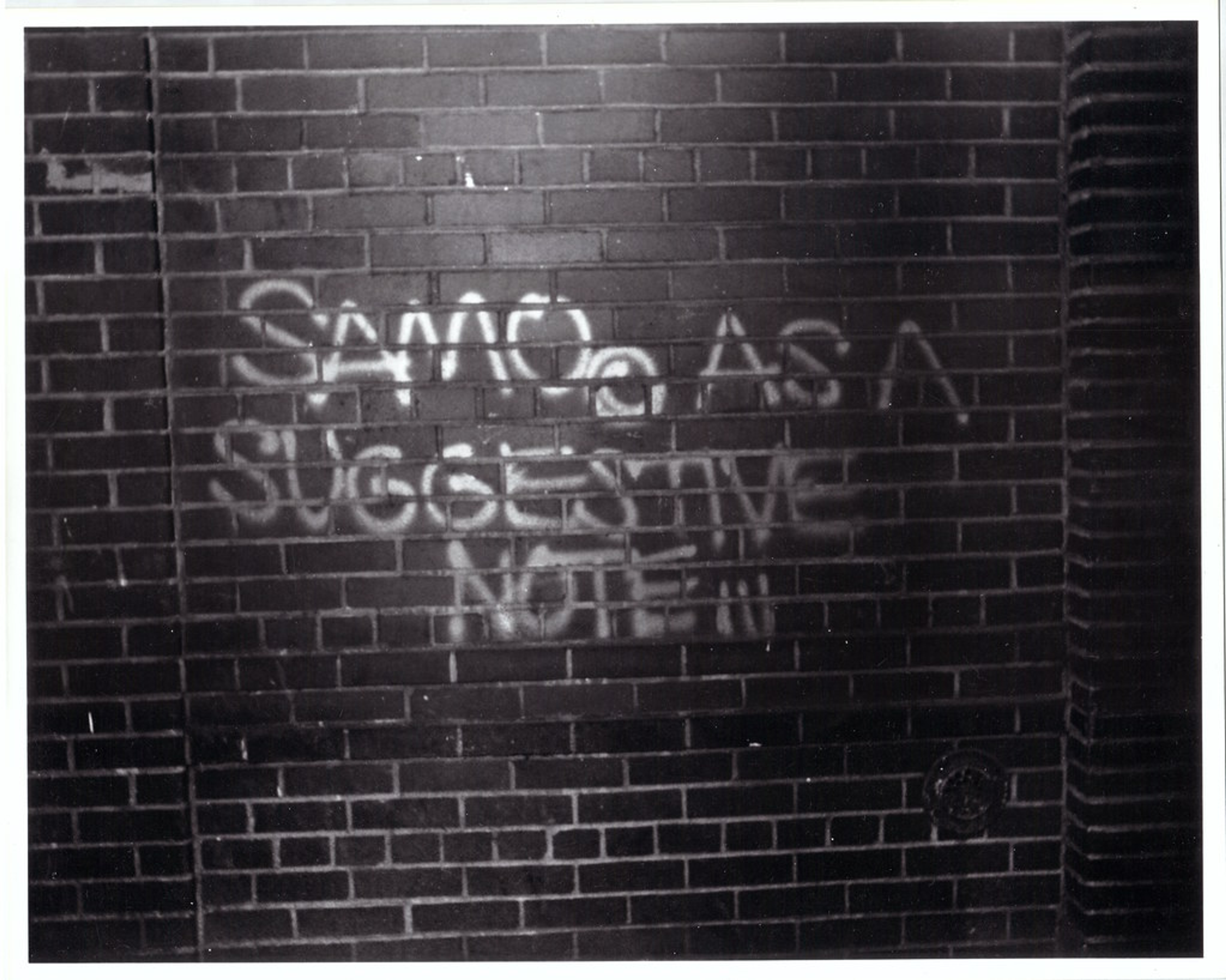 Une image d'un graffiti de SAMO sur un mur de briques. On peut y lire : « SAMO AS A SUGGESTIVE NOTE. »