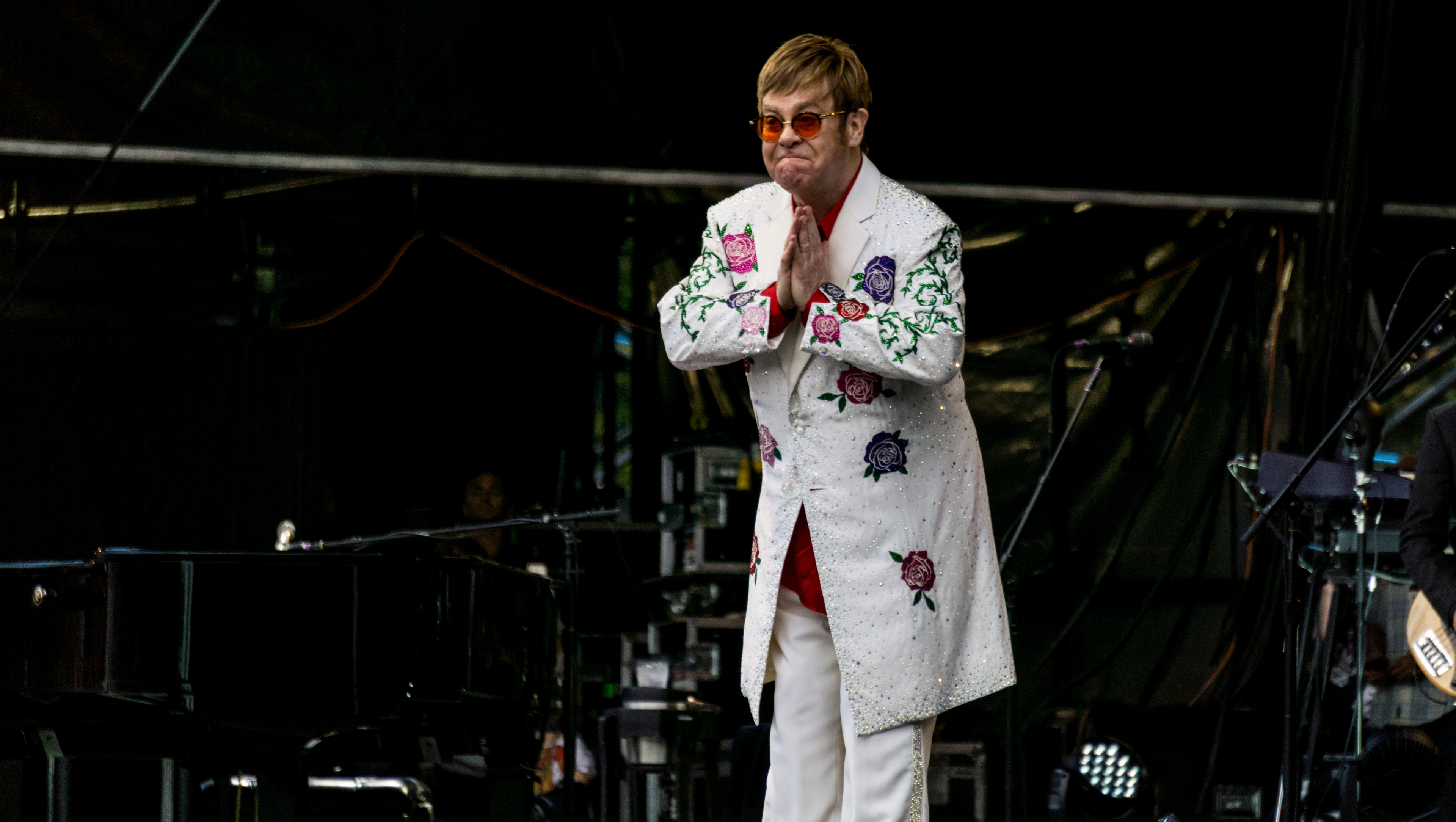 Cette photographie montre l'artiste Elton John sur scène, vêtu d'un costume blanc brodé de paillettes et de ses lunettes de soleil colorées emblématiques.