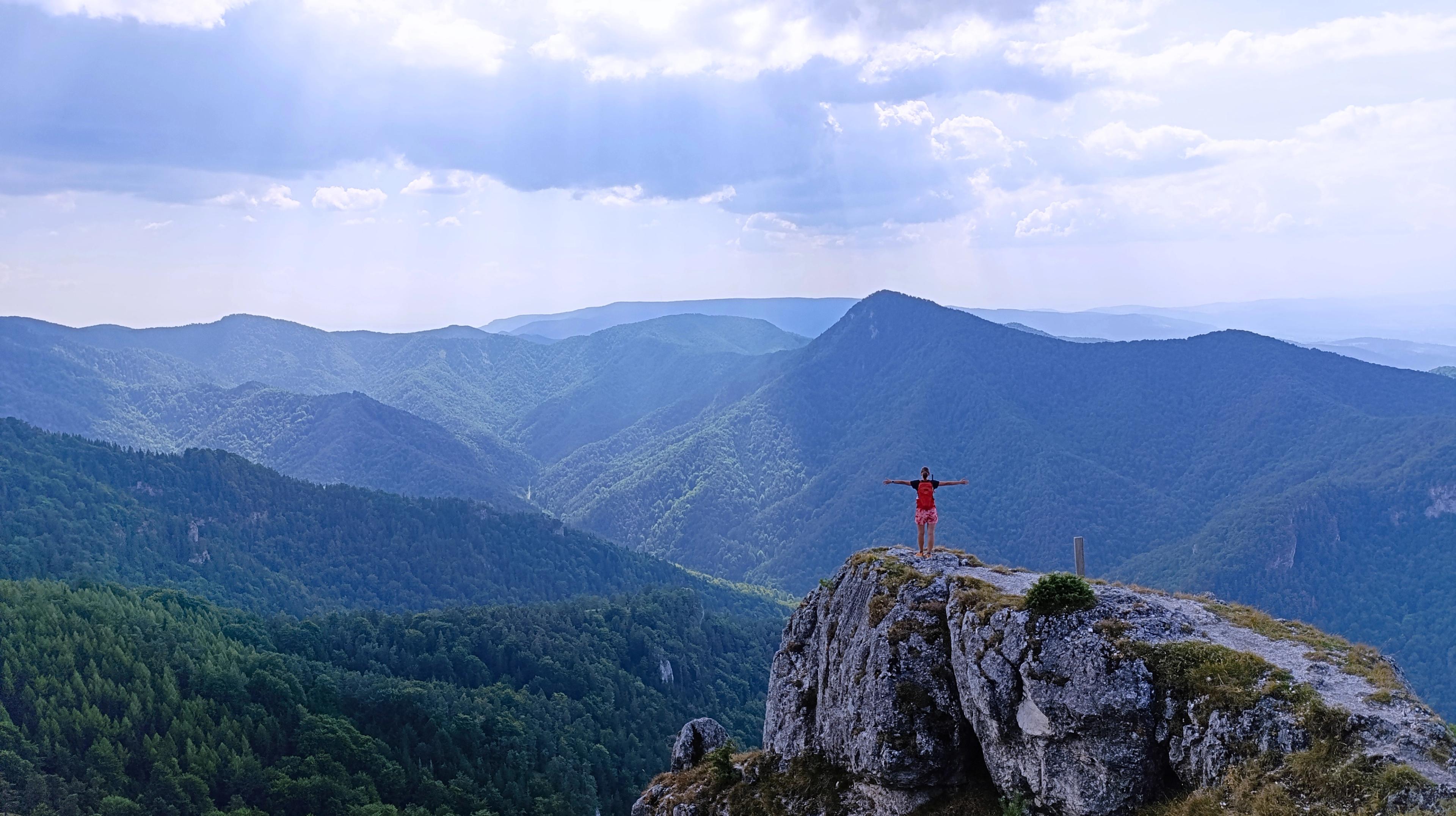 A lone figure standing on a rocky outcrop above wild forested ridges