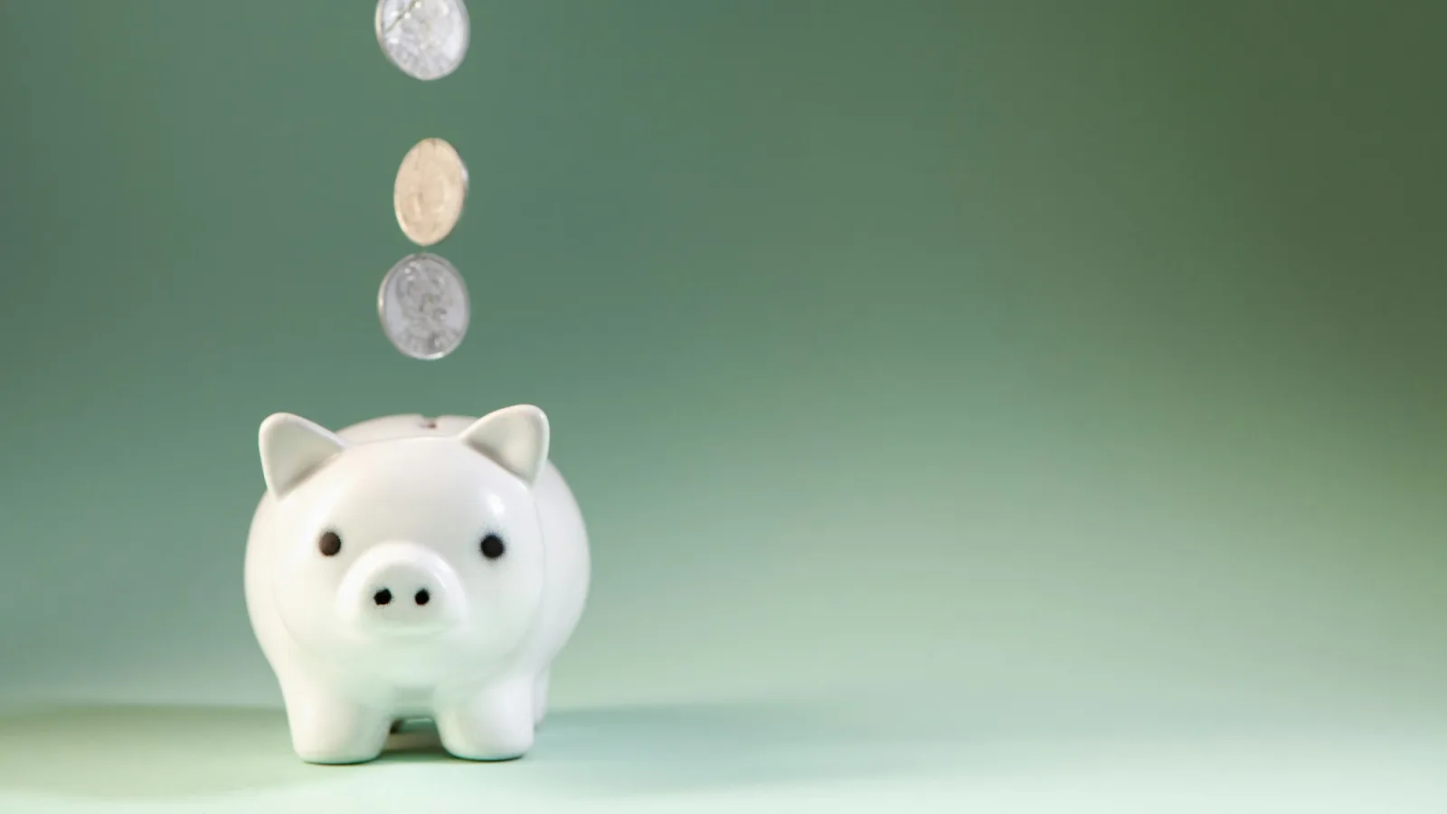 White ceramic piggy bank with three coins falling toward it against a soft green background