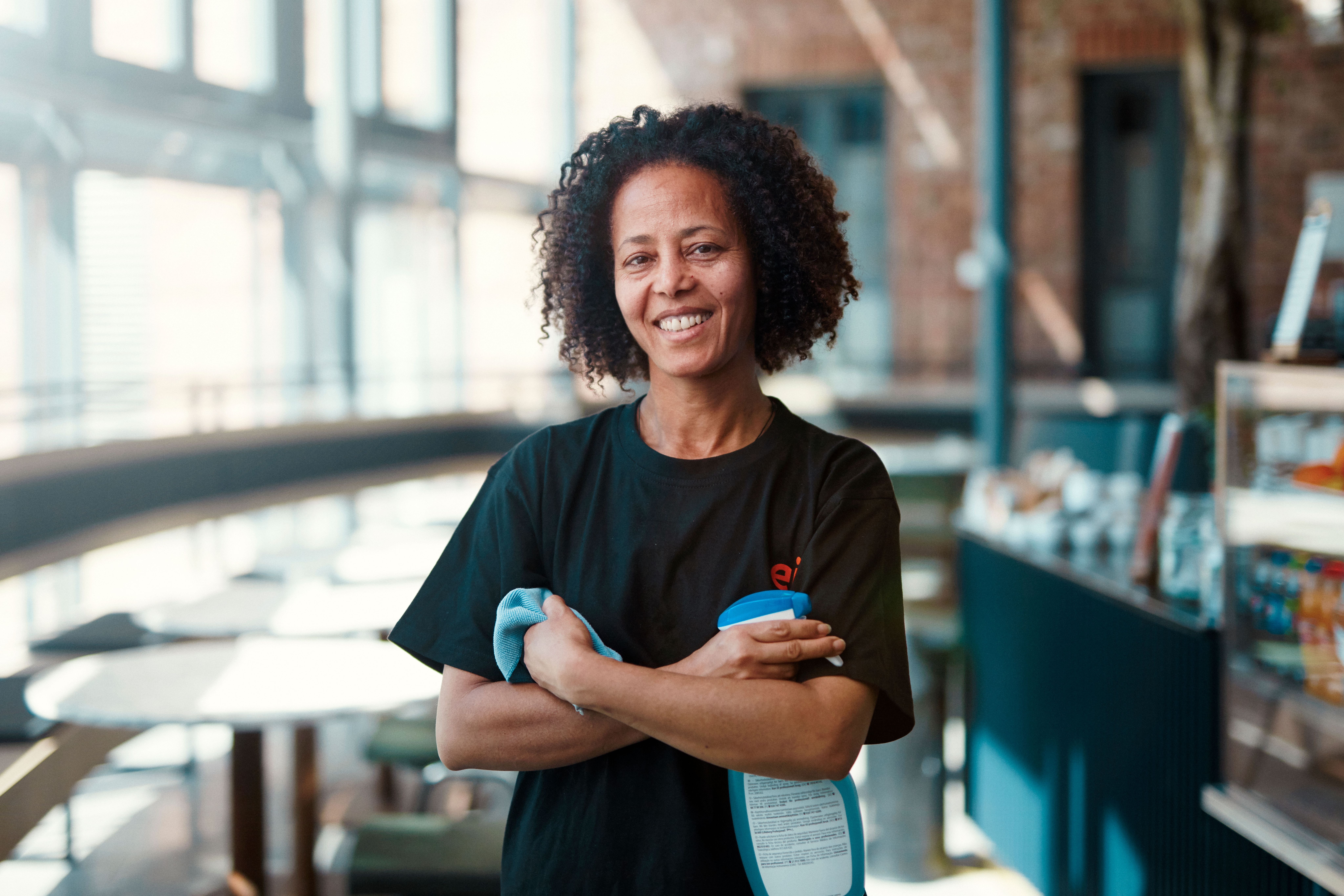 Bilde av dame fra Eir smiler med hendene i kryss. Hun har en klut i ene armen, og en sprayflaske med rengjøringsmiddel i den andre hånden