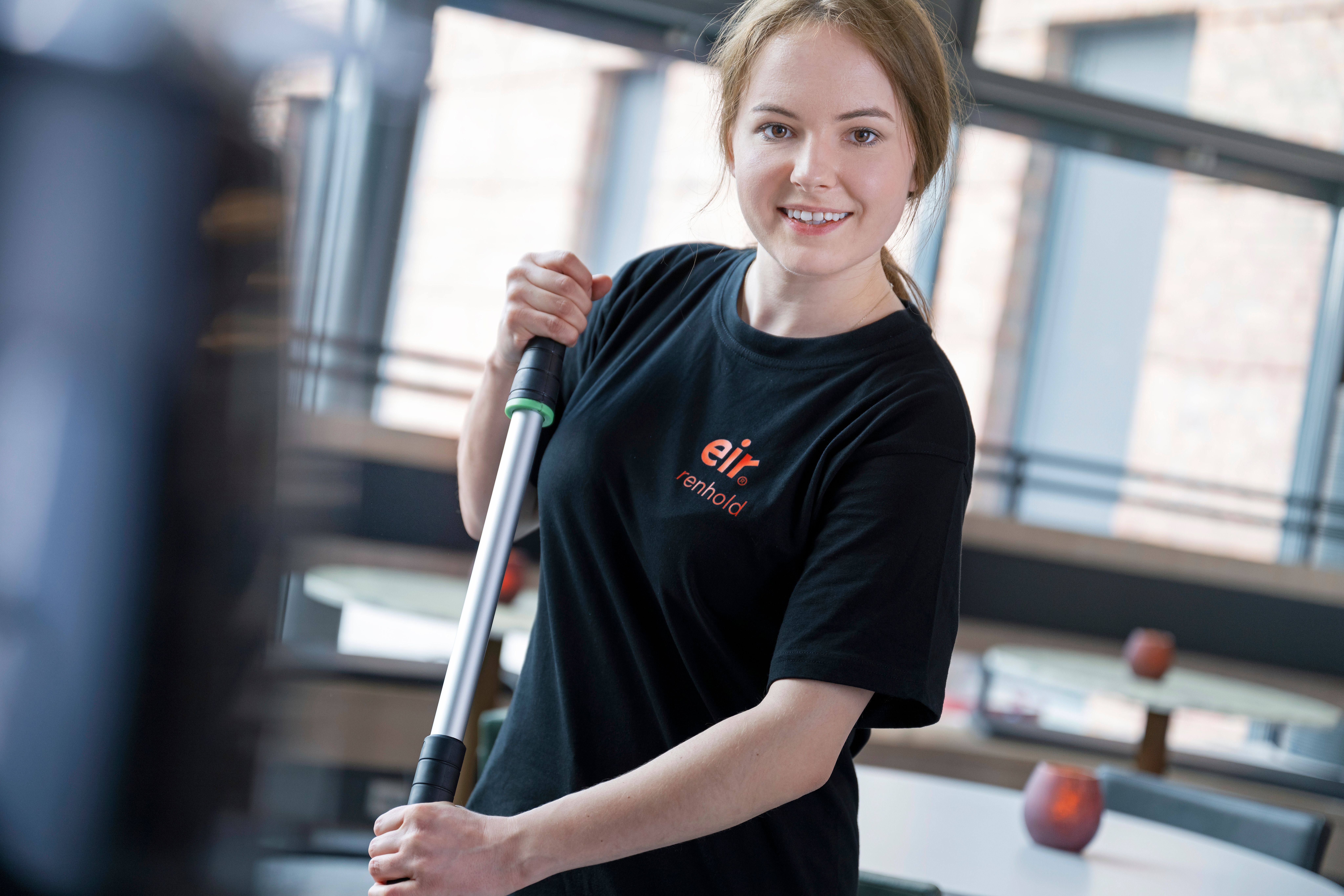 Foto av dame og mann i Eir-uniform står sammen i en kantine. Damen holder en i vasketralle og mannen holder en gulvmopp