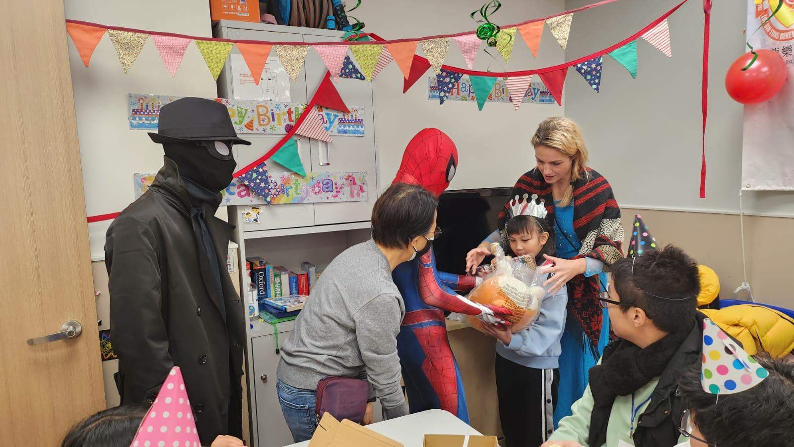 Adults and costumed performers help a child open a gift.