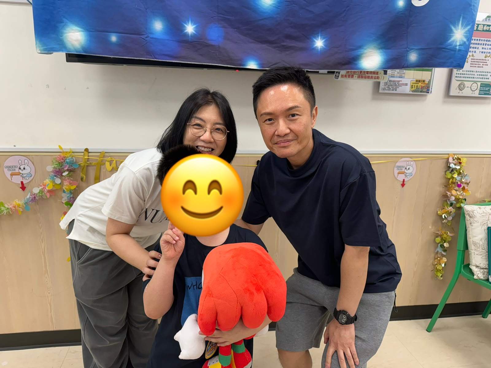 Two adults pose with a child holding a large red toy.