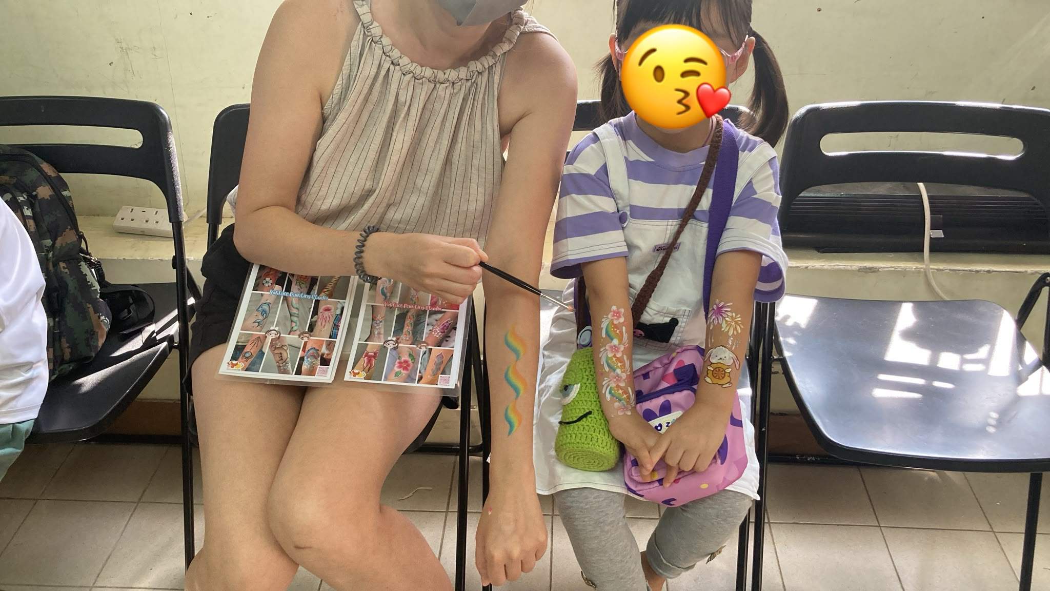 A face painter displays colorful designs beside a seated child.