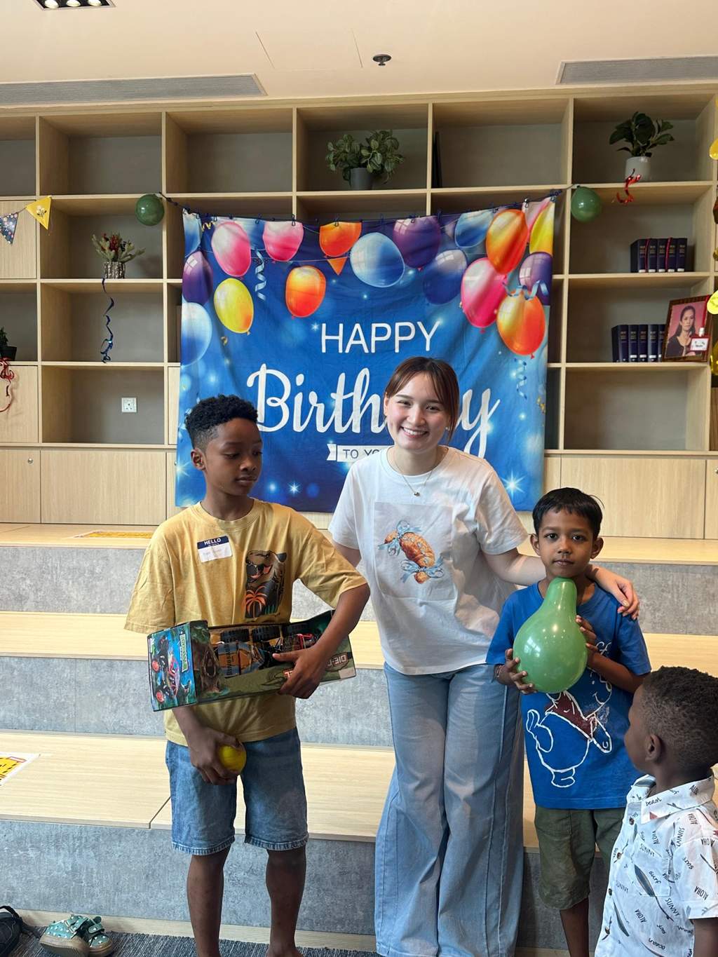 An adult stands between two children holding gifts and balloons.