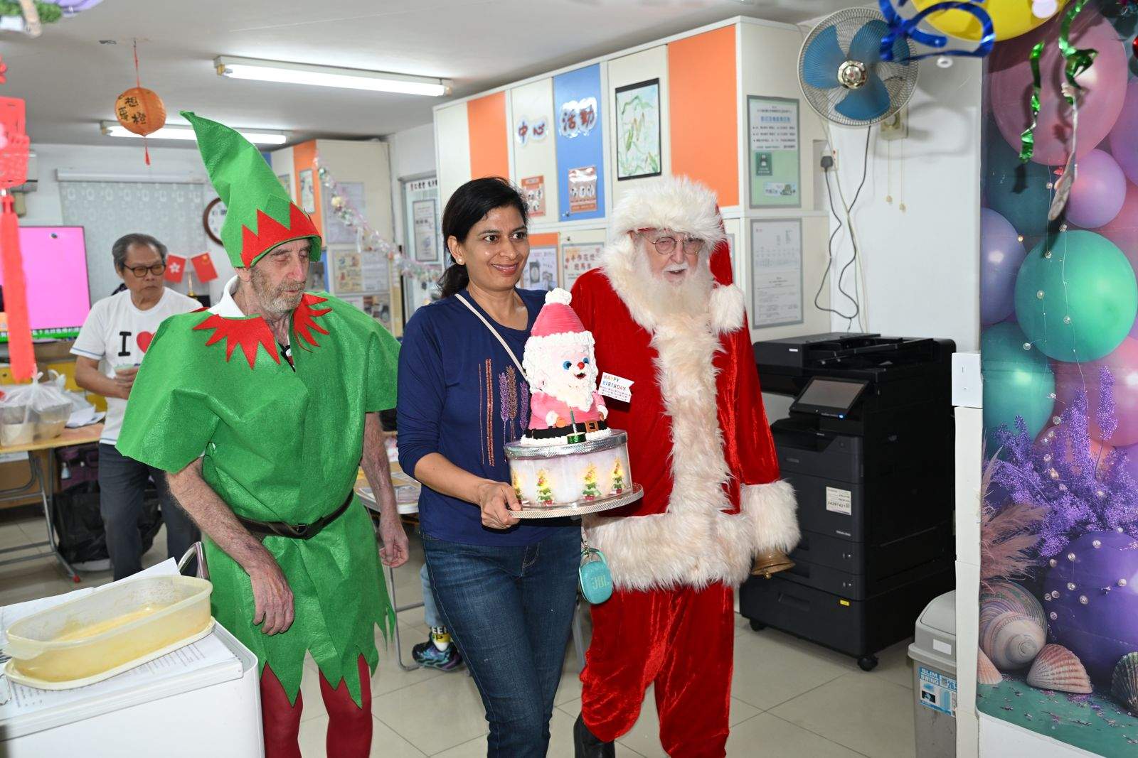 Two costumed adults stand beside an adult holding a cake.