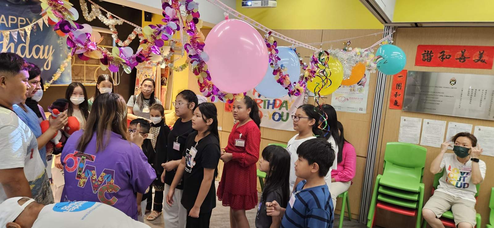 A crowded group watches an activity beneath colourful balloons.