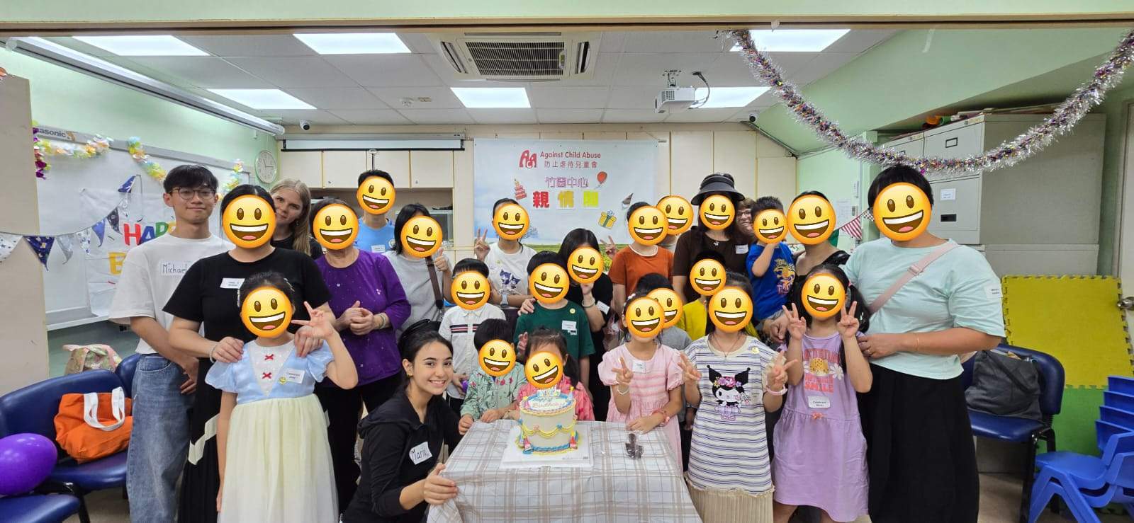 A large group gathers behind a decorated cake table.