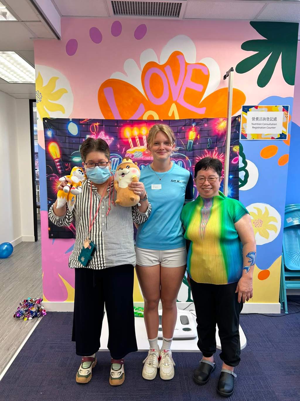 Three people pose with stuffed toys before a colourful backdrop.