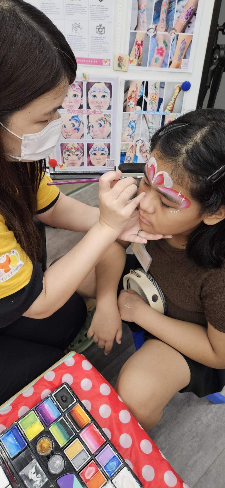 An adult paints a seated child's face at a small table.