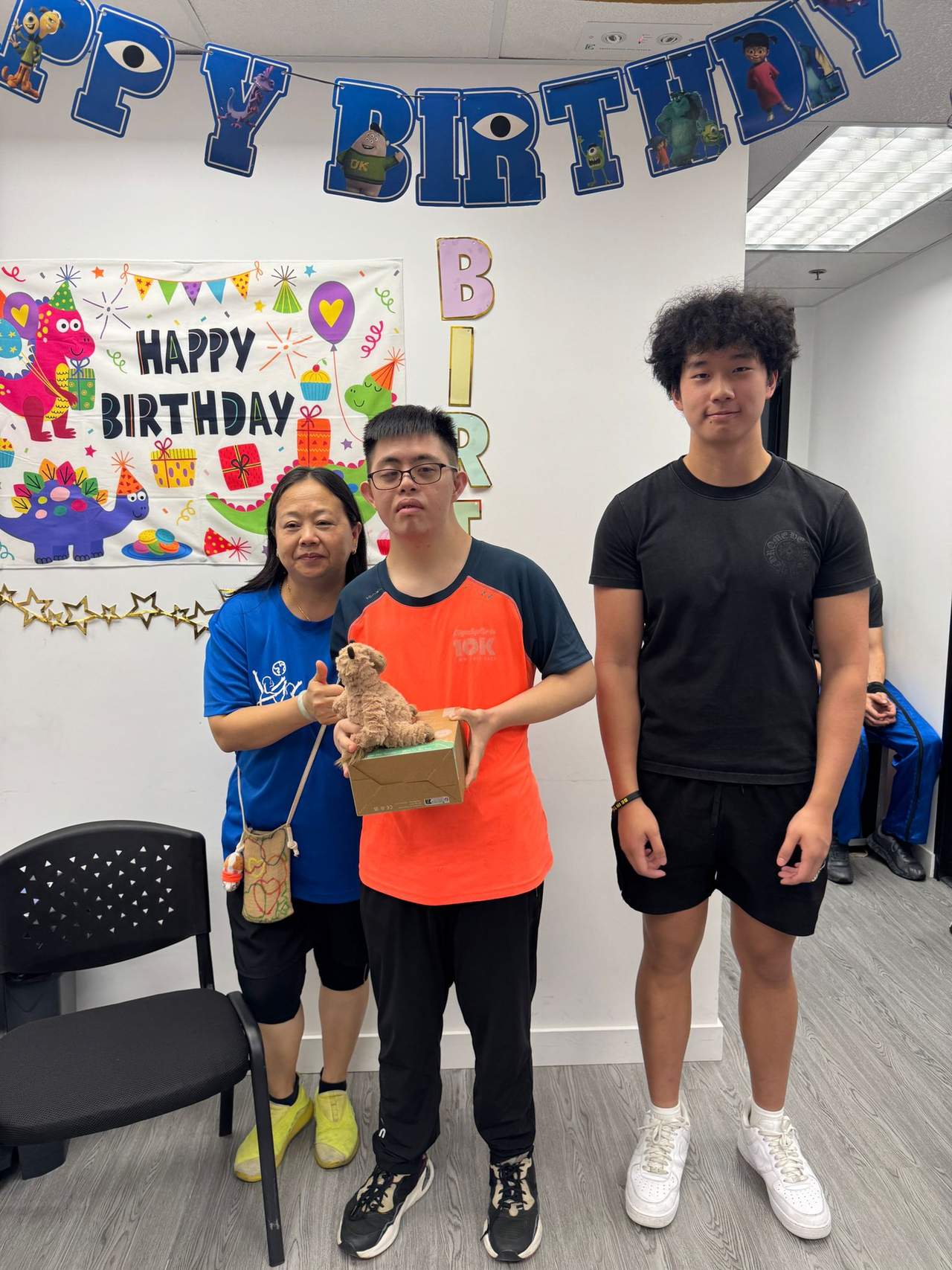 Three people stand together with a gift beneath birthday decorations.