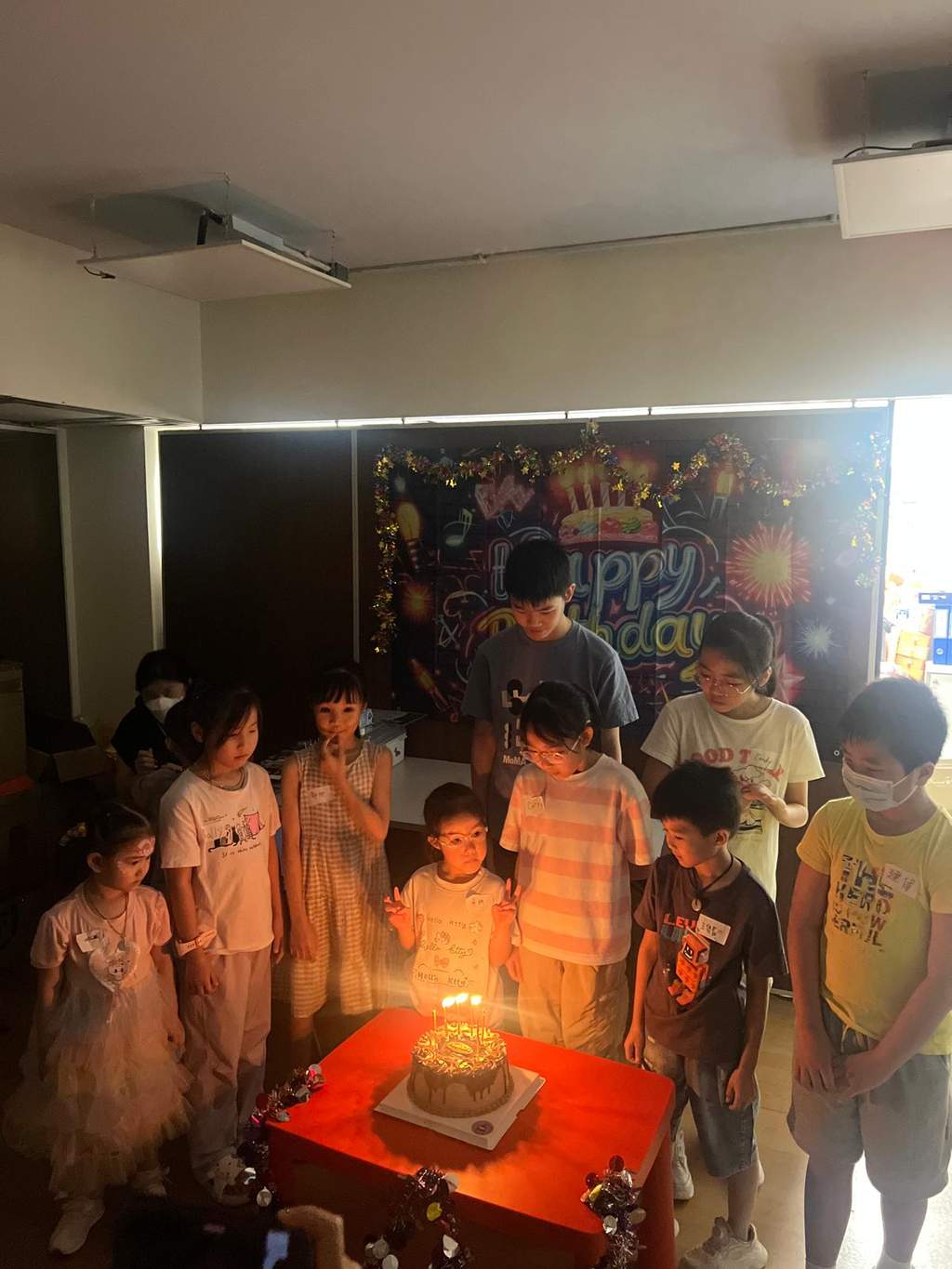 Children stand around a candlelit cake in a darkened room.