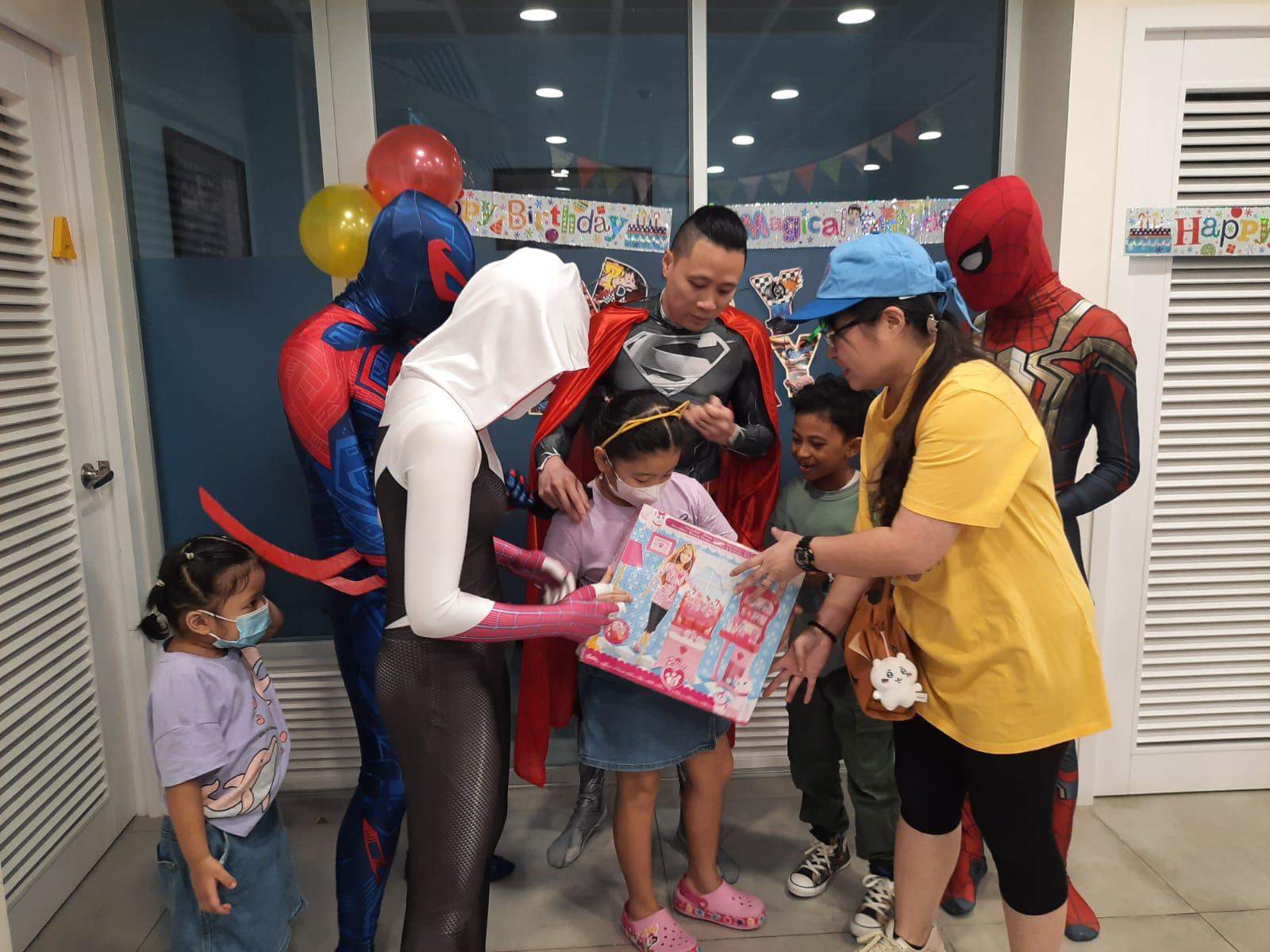 Costumed performers and an adult help a child hold a boxed gift.