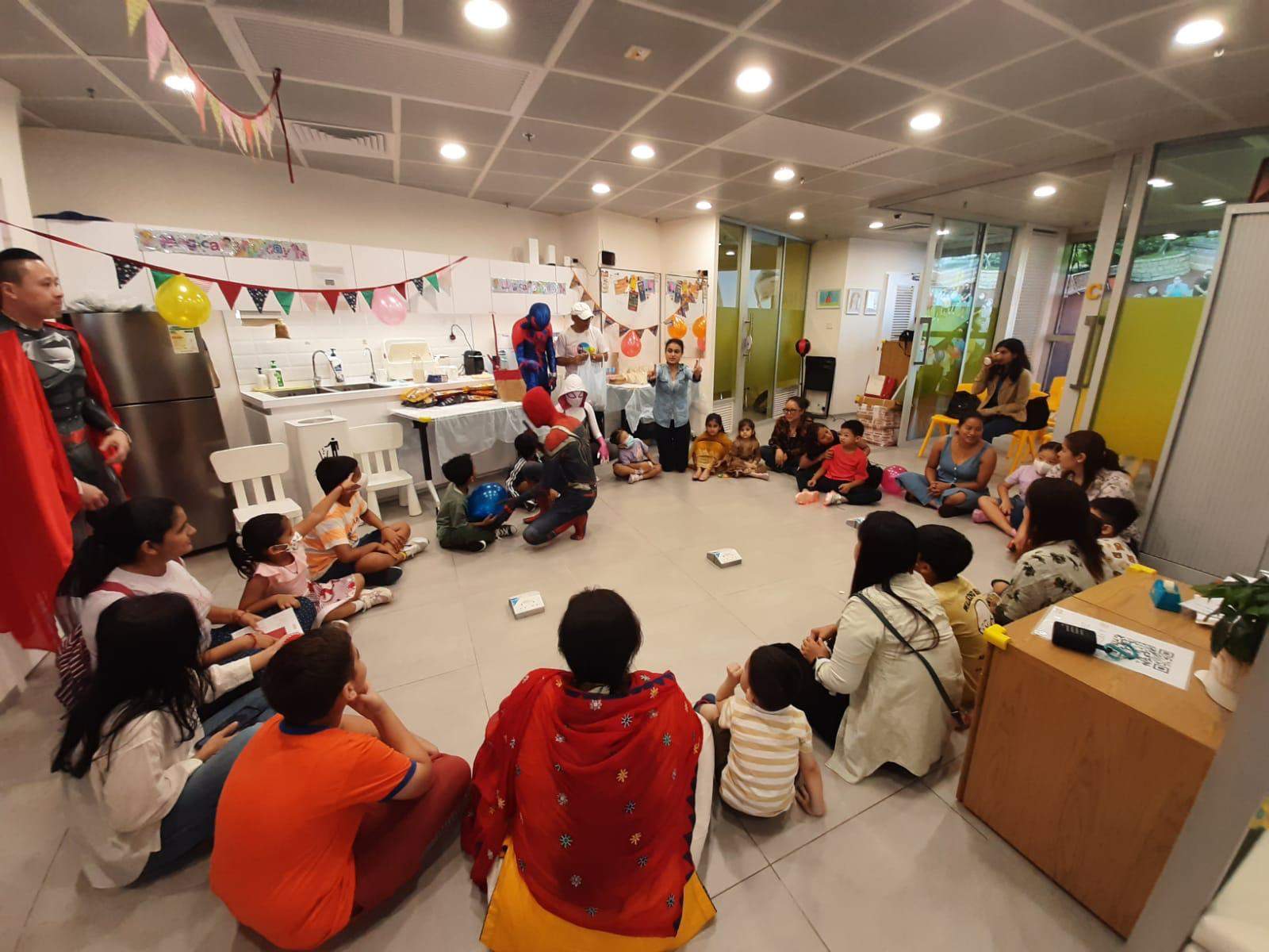 Adults and children sit in a wide circle around the room.