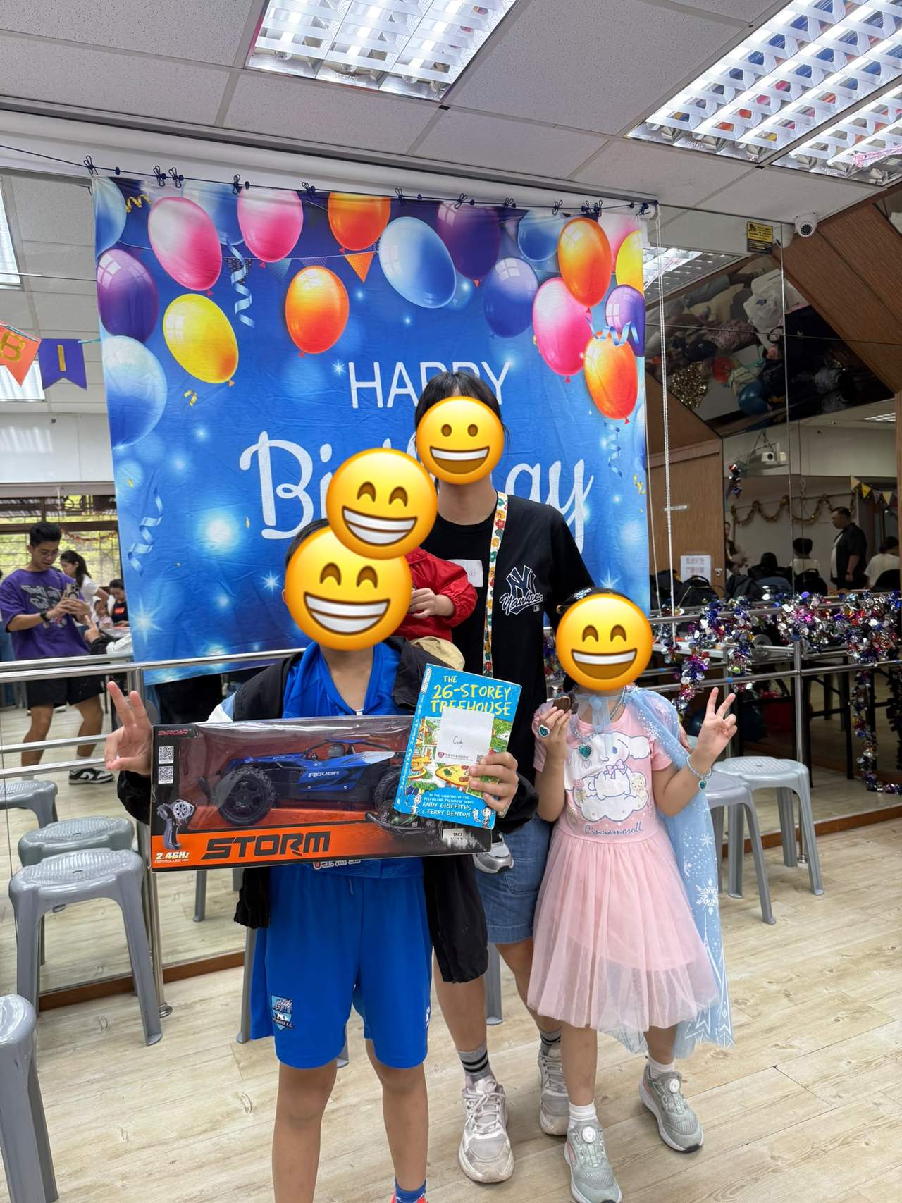 Two children display large boxed gifts before a balloon backdrop.