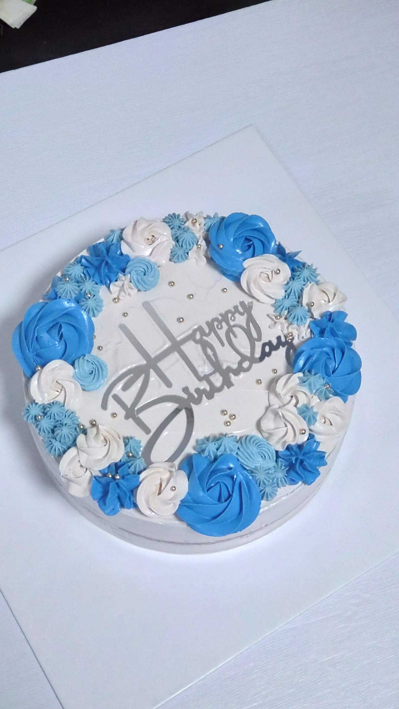 A white birthday cake ringed with blue and white buttercream rosettes, by Bakelicious By Aisha.