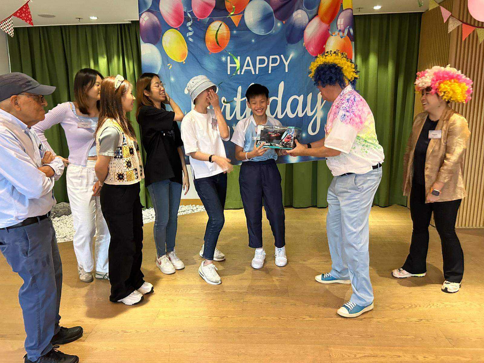 Adults present gifts to children near the birthday backdrop.