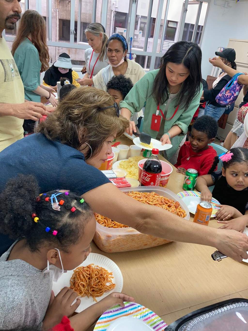 Adults serve plates of noodles to children around a crowded table.