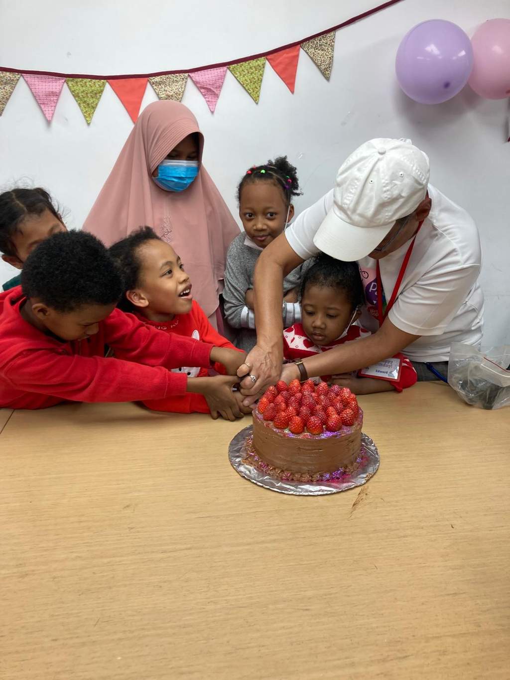 Children and adults reach together towards a strawberry-topped cake.