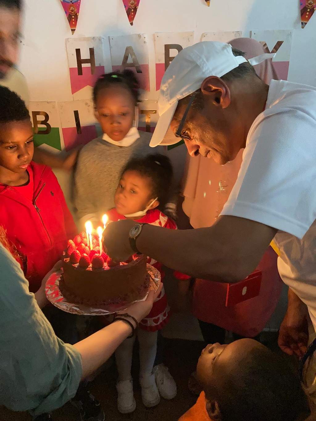 Adults hold a candlelit cake as children gather closely around.