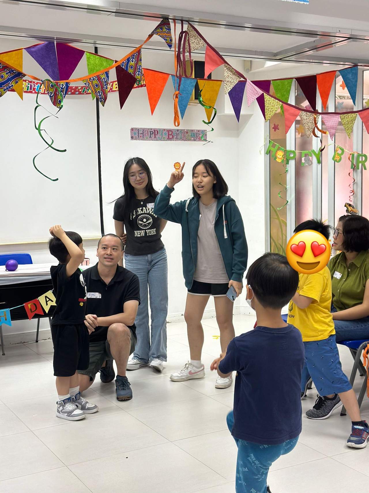 An adult gestures while children watch from blue chairs.