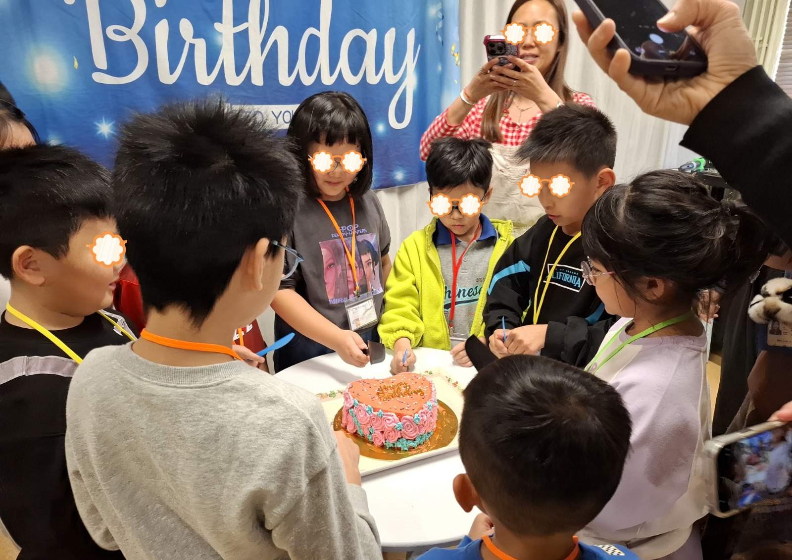 Children gather closely around a cake while adults take photographs.
