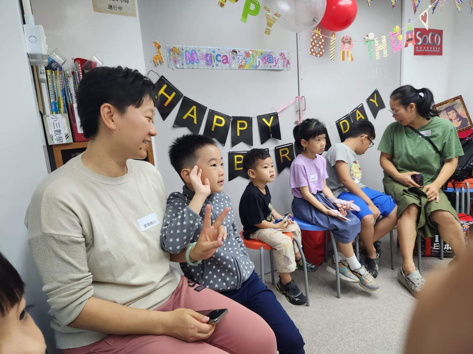 Adults and children sit along a wall beneath birthday bunting.