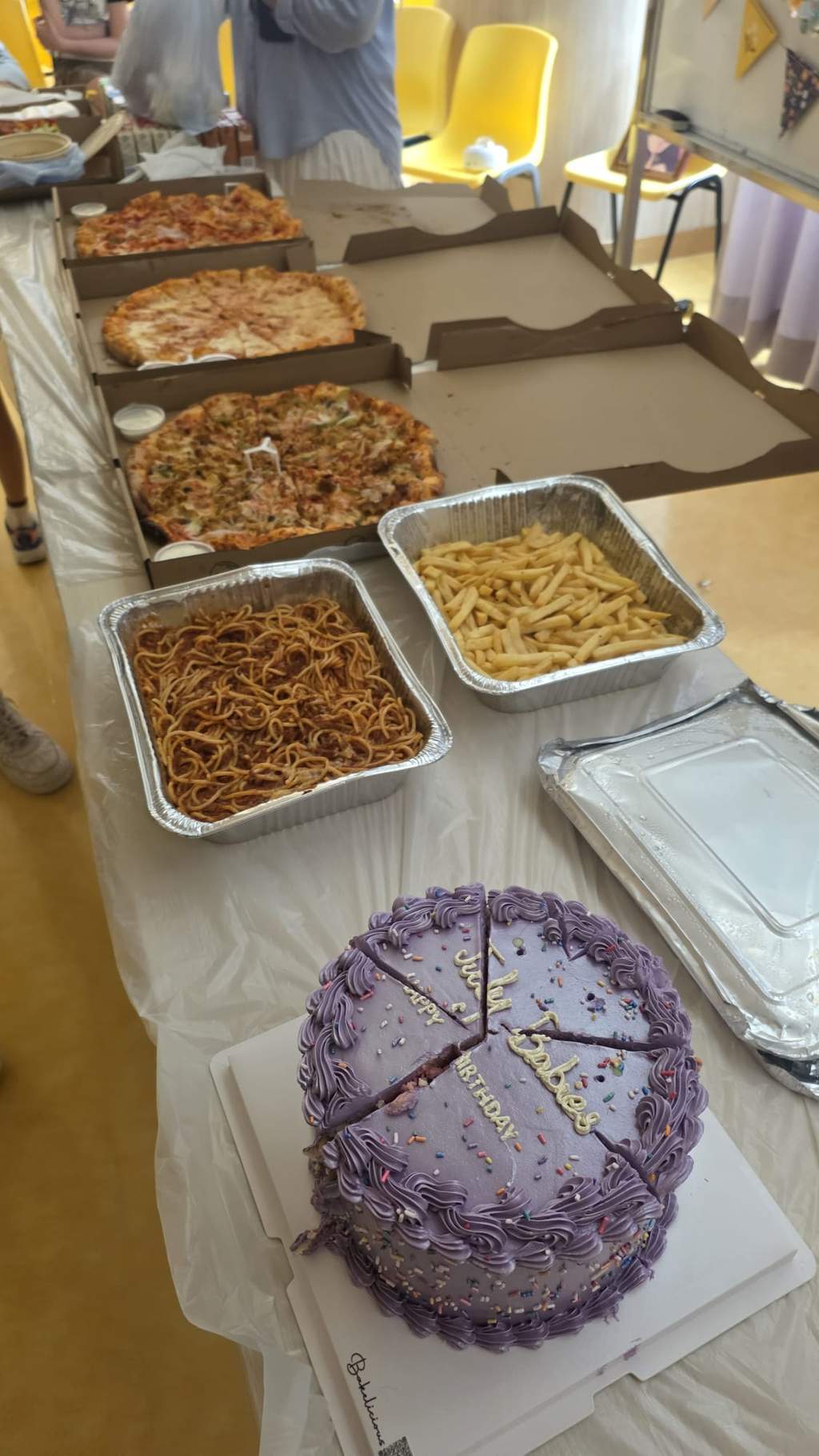 Pizzas, noodles, fries, and sliced cake are arranged on a table.