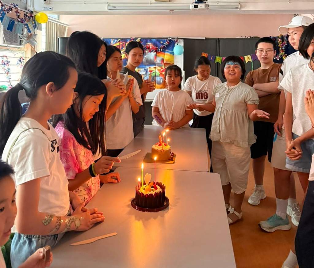 Children and adults gather around two candlelit cakes on a table.
