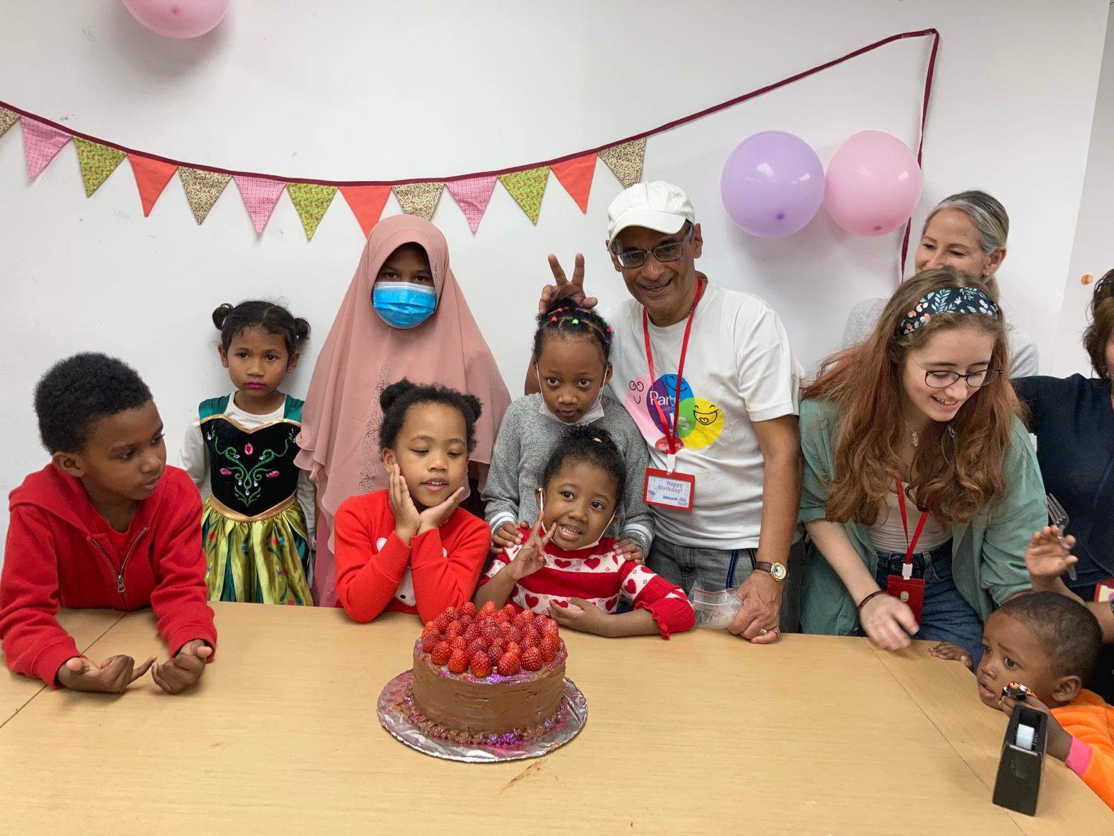 Children and adults gather behind a strawberry-topped cake on a table.