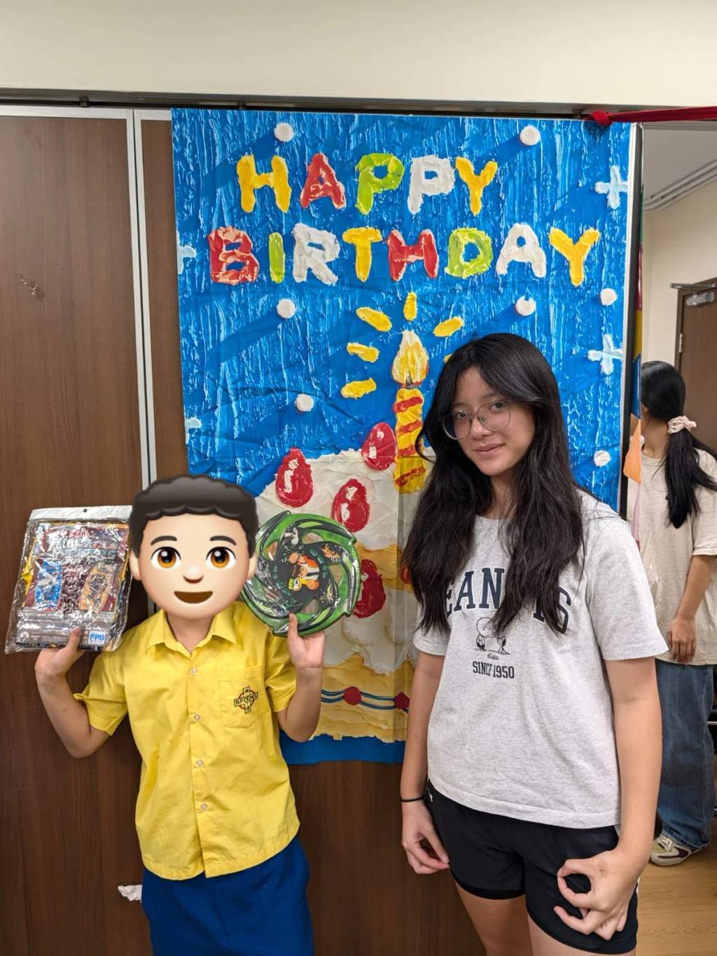 A child displays party gifts beside a volunteer and birthday backdrop.
