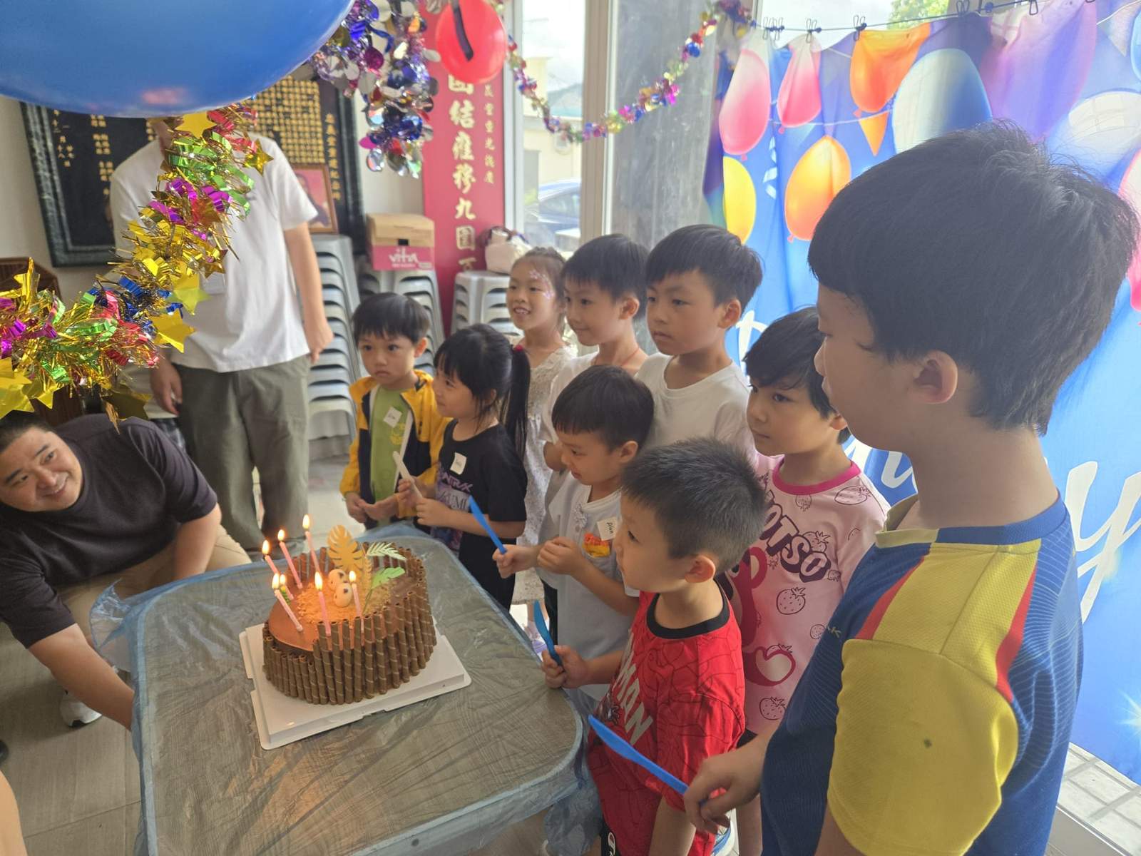 Children gather around a candlelit cake on a table.