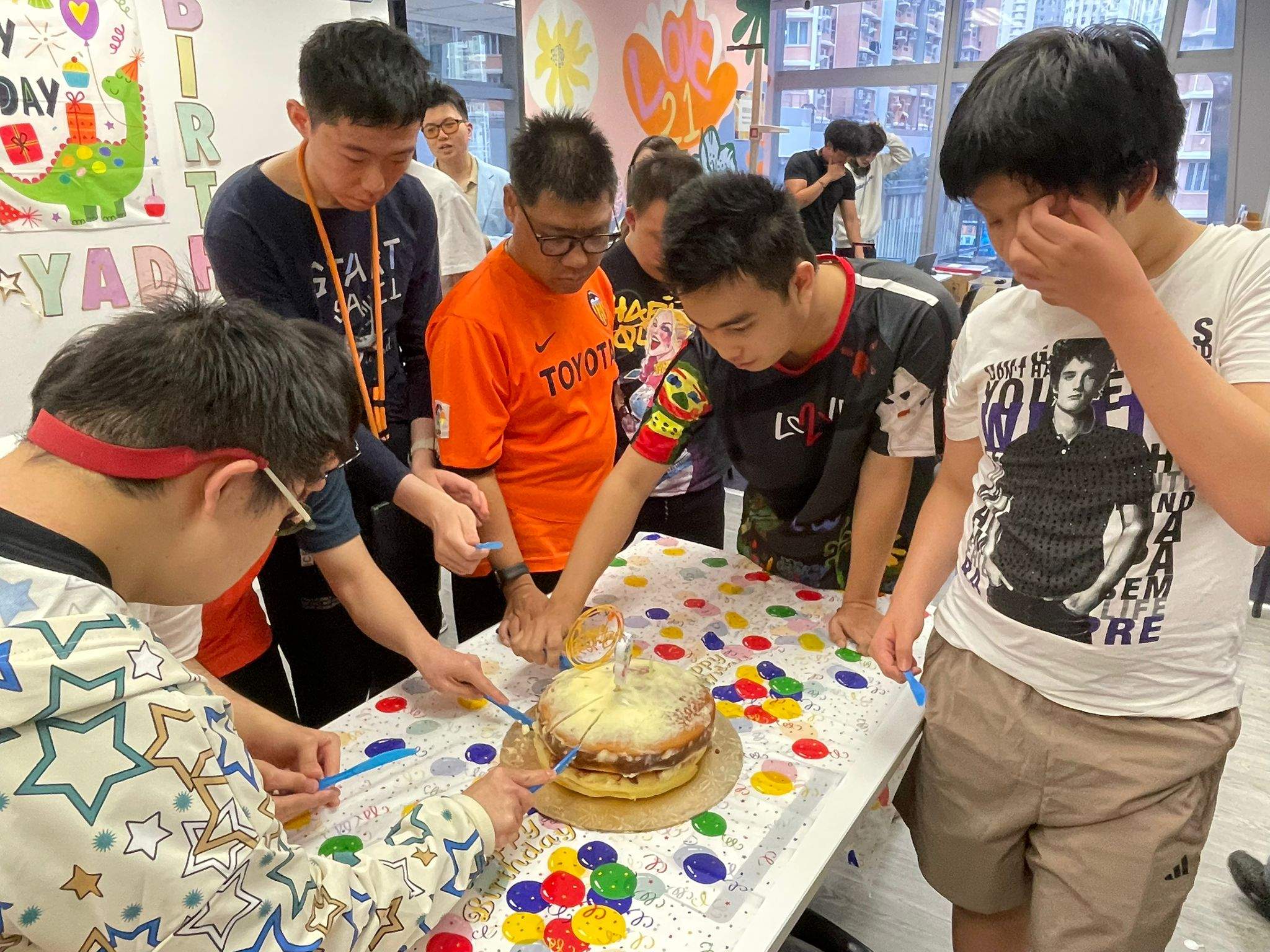 Several people gather around a cake decorated with colourful pieces.