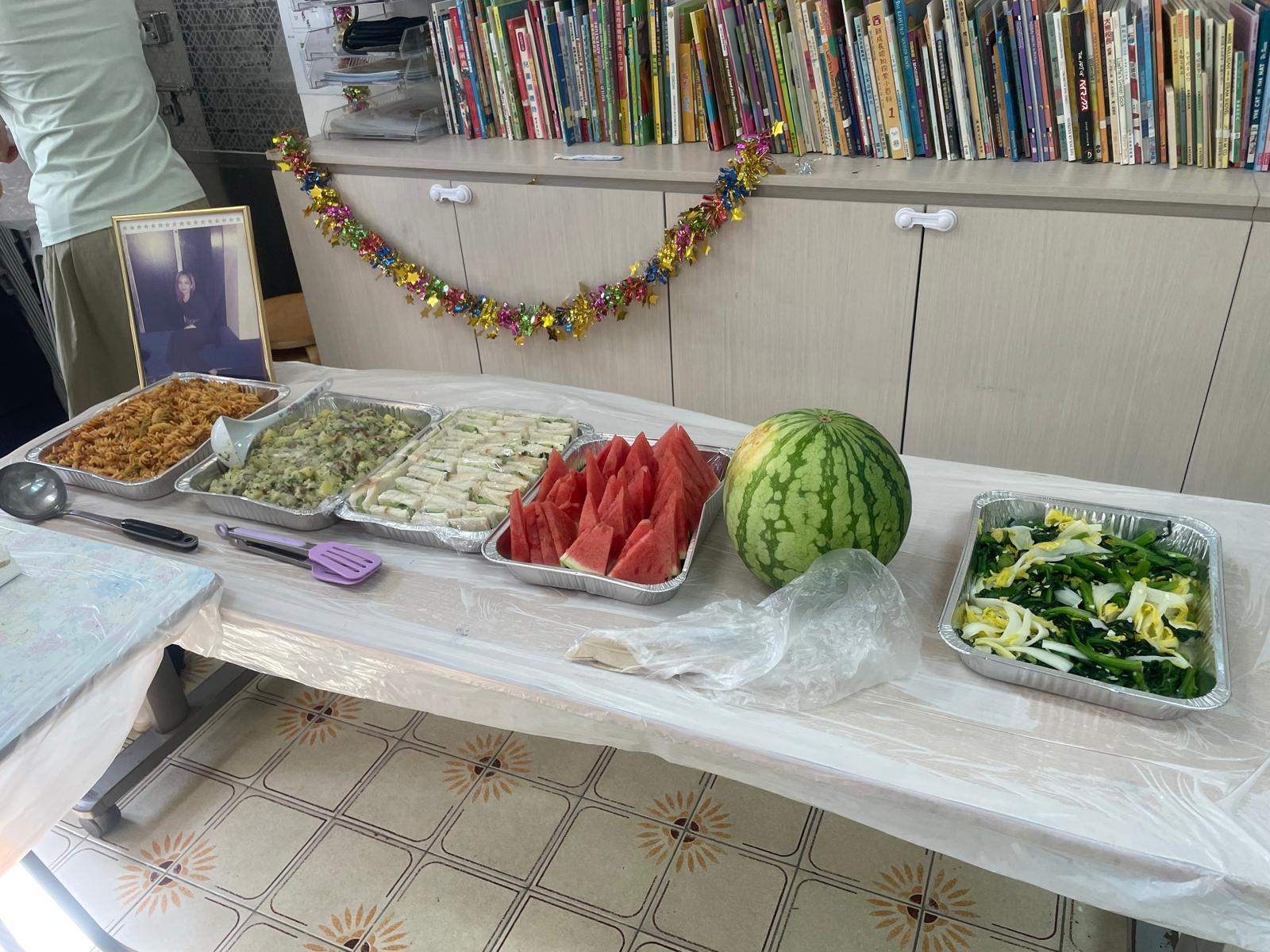 A long table displays fruit, noodles, vegetables, and other dishes.
