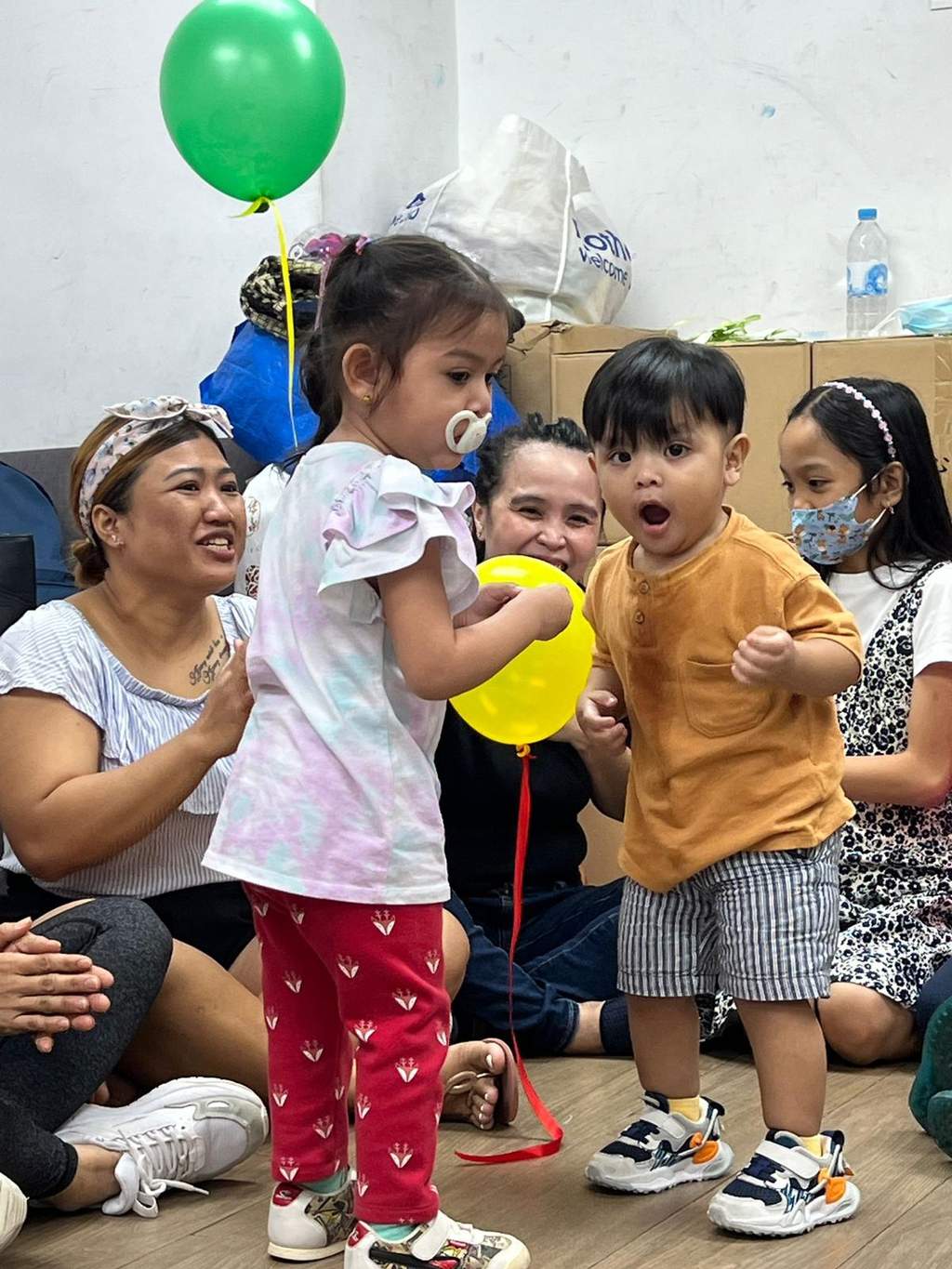 Children hold balloons together as seated adults gather behind them.